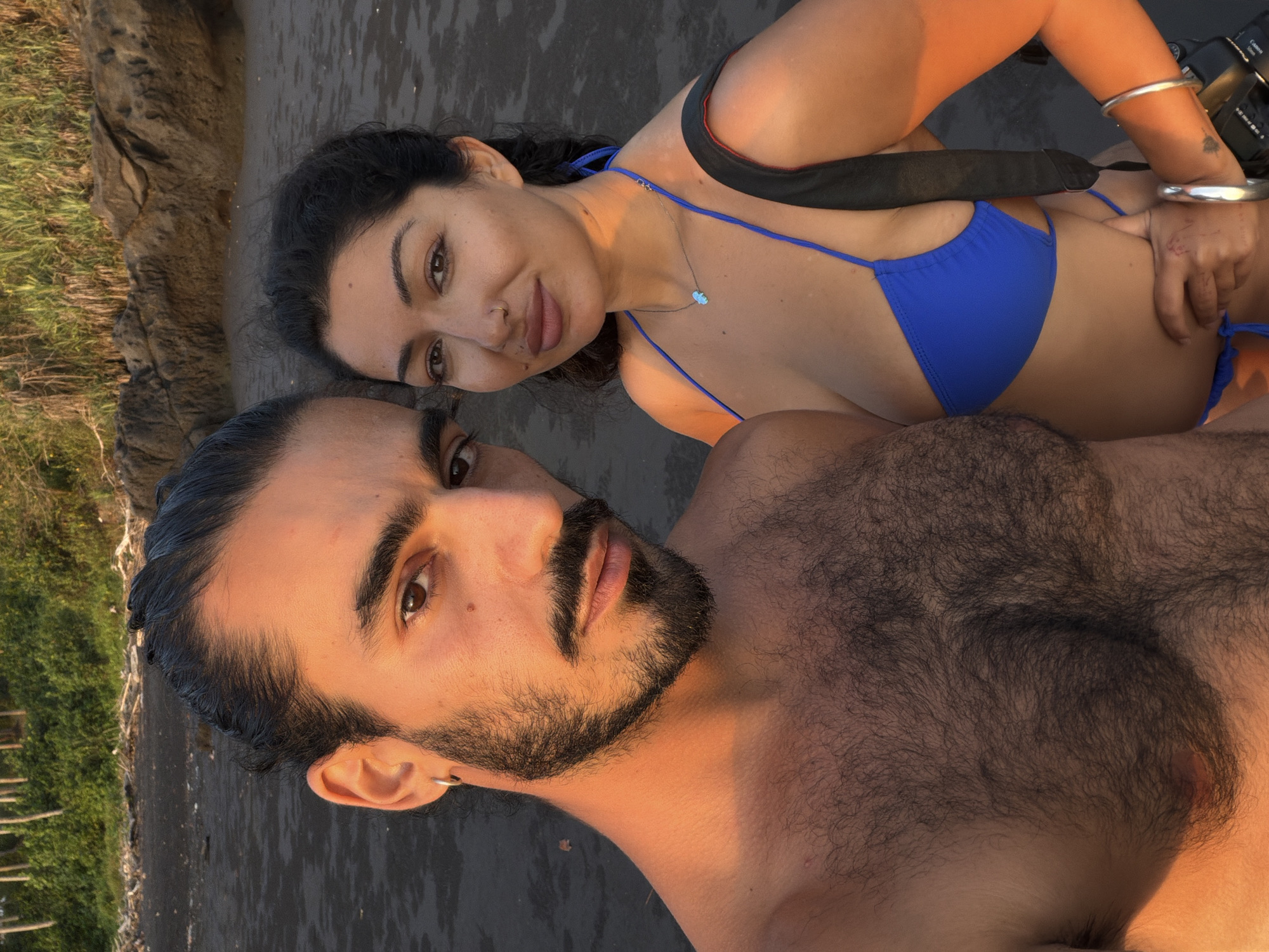Two individuals, a man and a woman, are posing for a selfie on a beach with dark sand. The man, on the left, is shirtless and has dark hair pulled back, a beard, and a mustache. He is wearing a silver hoop earring. The woman, on the right, is wearing a bright blue bikini and a silver necklace with a light-colored pendant. She has dark curly hair and a nose ring. A black strap is visible across her shoulder, suggesting she is carrying a camera bag. In the background, there is lush green foliage, rocky outcrops, and the ocean. The setting sun casts a warm, golden light on the scene. The location is identified as Selemadeg, Indonesia.