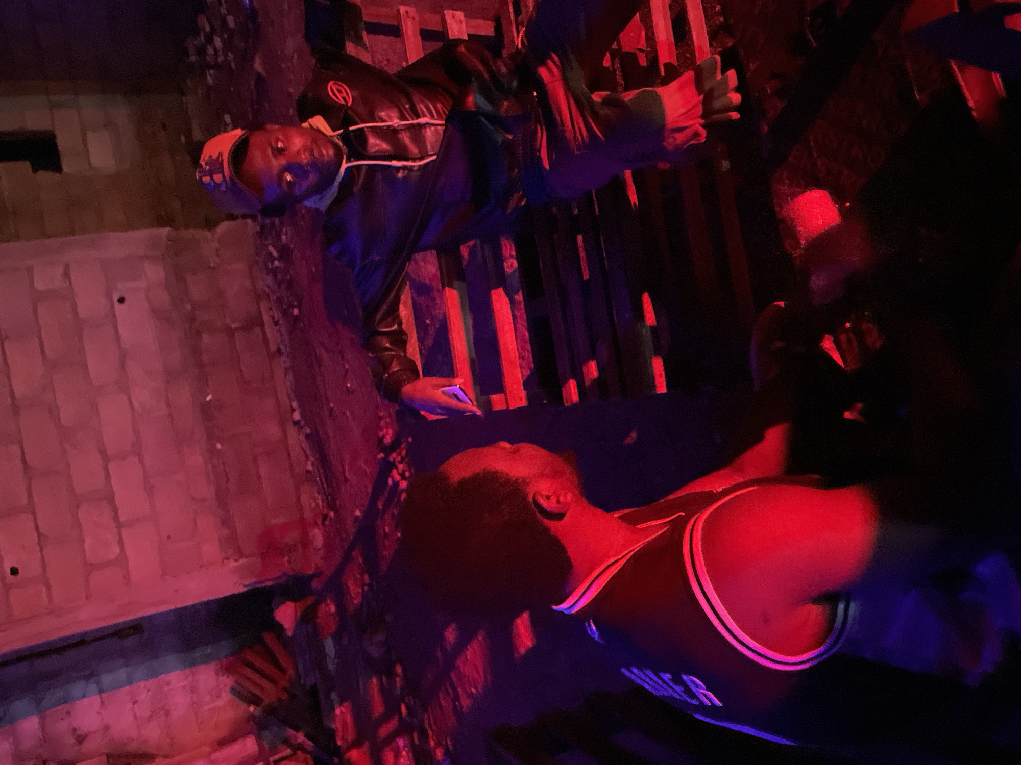 In Durimi, Nigeria, two young men are seated on a wooden bench in what appears to be an outdoor urban setting at night. The scene is illuminated by red and purple lights, creating a dramatic and somewhat disorienting atmosphere. The ground is littered with debris, suggesting an unfinished or neglected area.
The man on the right is wearing a dark jacket with a hood and a baseball cap. His arms are resting on the back of the bench, and he is looking towards the left. The man on the left is wearing a dark sleeveless jersey with red and white trim. He has dark hair and is facing away from the camera, looking towards the man on the right. His left arm is resting on the bench, and he appears to be engaged in conversation or observation of the other individual.
The background consists of a brick wall, partially illuminated by the colored lights. The overall impression is one of a candid moment captured in a dimly lit, possibly informal public space in the city of Durimi.