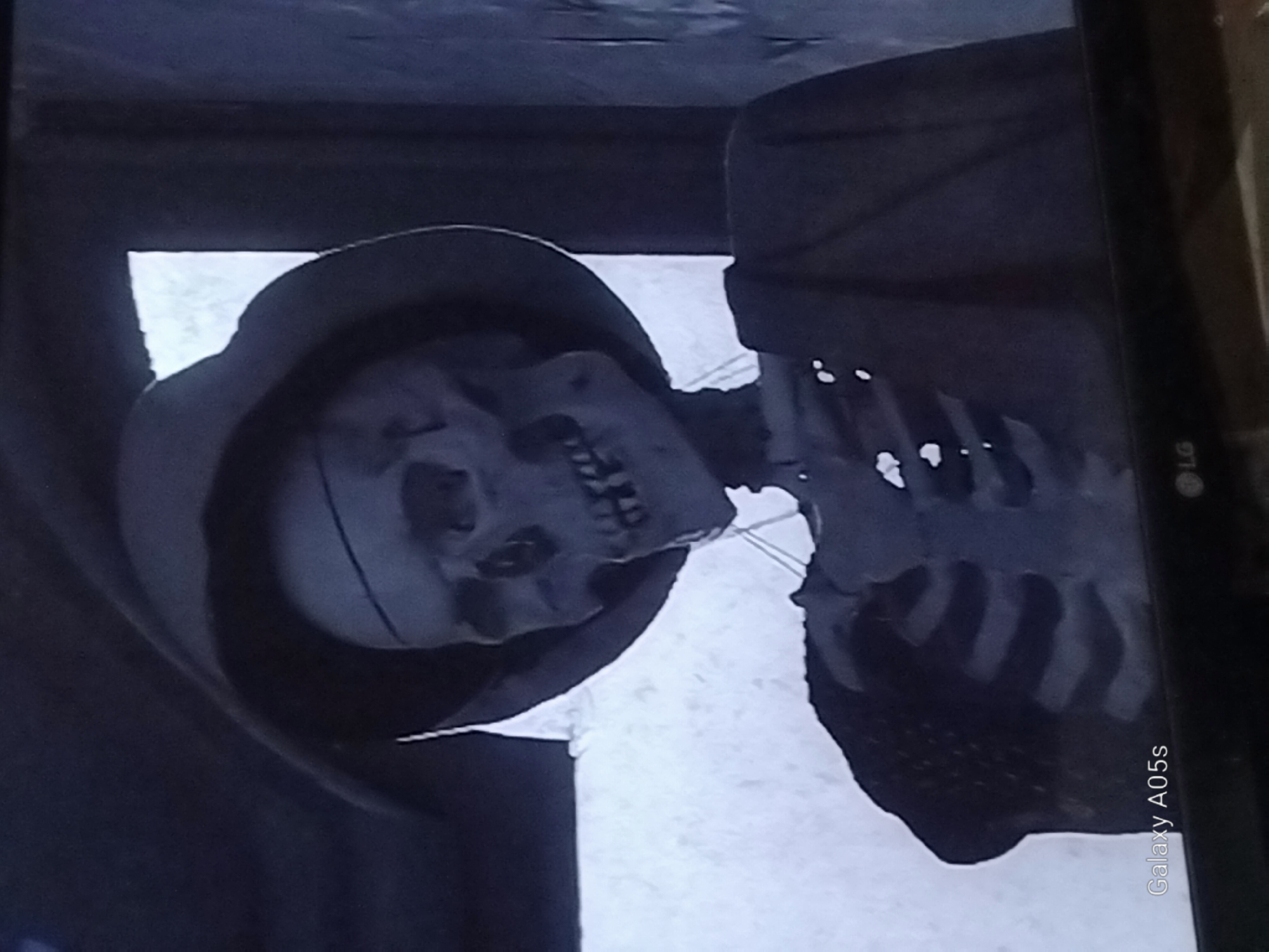 The image shows a close-up view of a skeleton wearing a hat and dark clothing. The skeleton is positioned in what appears to be an indoor setting, with a plain background. The lighting is dim, creating shadows on the skeleton's face and body. There are no people visible in the image, and no explicit actions or interactions are depicted. The context of its location in the city of Druzhba, Russia, is not evident from the visual information provided in the image.