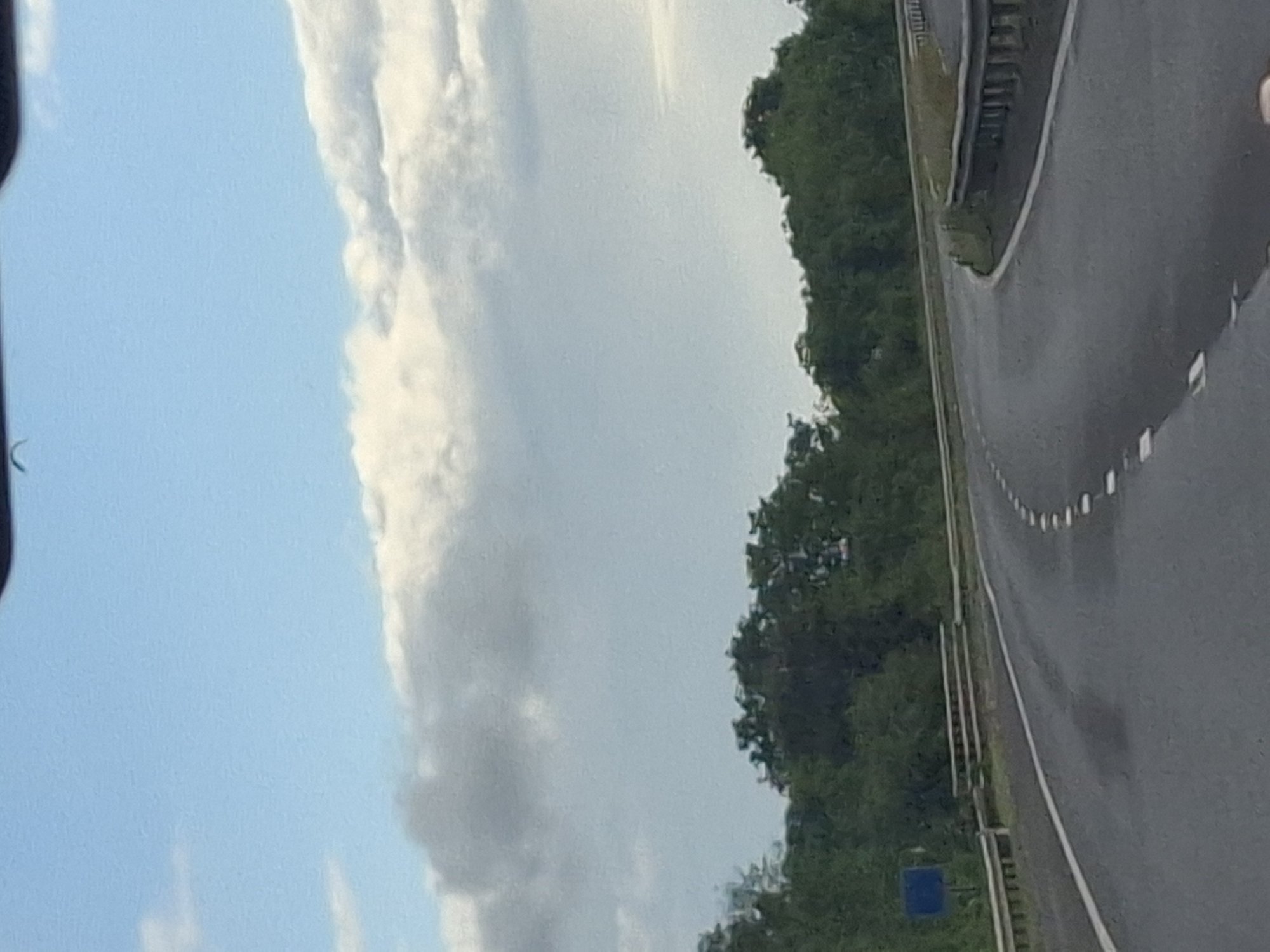 The image shows a view from a moving vehicle on a highway in Darlington, United Kingdom. The road curves to the right, with white dashed lines marking the lanes. Guardrails are visible on both sides of the road, with a blue sign on the left side. Dense green trees line the background, and the sky above is a pale blue with scattered white and gray clouds. The overall scene depicts a typical highway landscape.