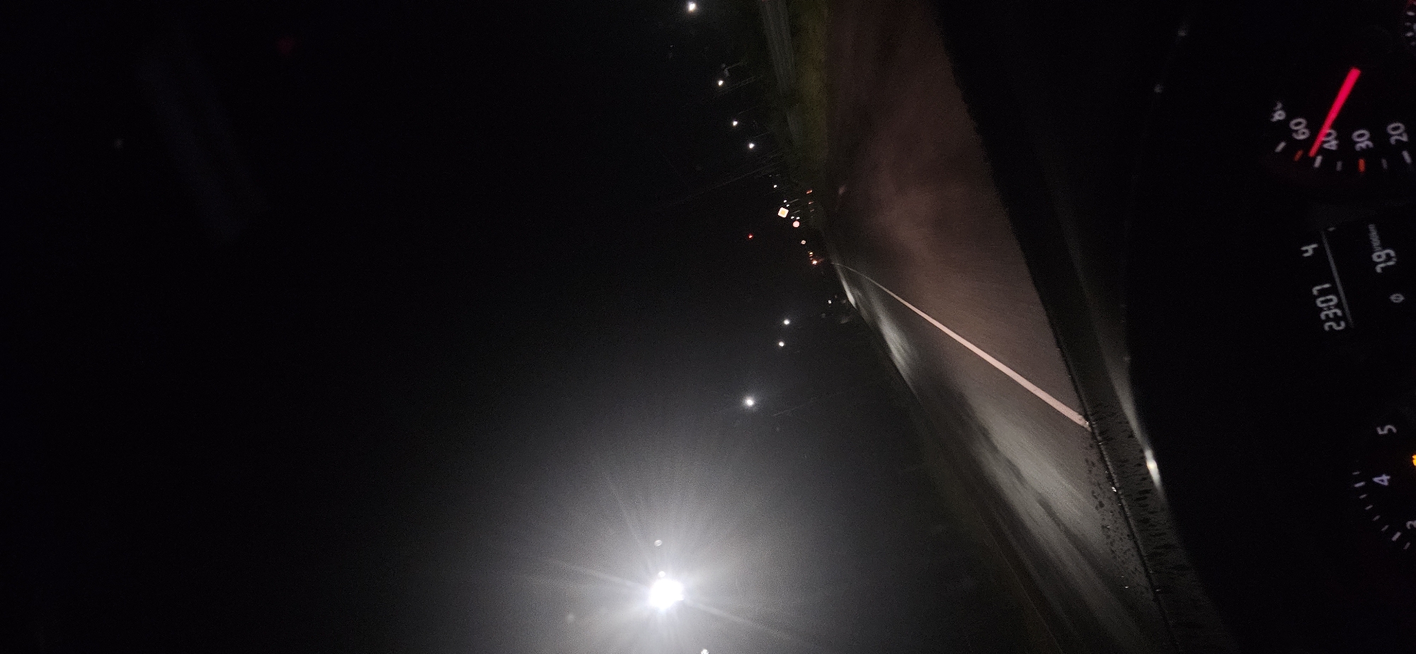 A forensic image analysis reveals a nighttime scene captured from the interior of a moving vehicle, looking forward through the windshield onto a wet, two-lane asphalt road. The road, identifiable by a continuous white dividing line, recedes into the distance and shows reflections from overhead illumination, indicating a wet surface. Streetlights line both sides of the road, with a prominent, bright light source on the far left generating significant lens flare. Faint red lights are visible further down the road. The vehicle's dashboard occupies the lower portion of the image, displaying a digital clock at "23:07" and a fuel efficiency reading of "7.9 l/100km". A speedometer on the right features a red needle indicating a speed between 40 and 50 (likely km/h). A yellow warning light is illuminated on the lower left of the dashboard. The external environment is dark, typical of a rural or suburban road at night in the specified location of Momina Klisura, Bulgaria. No individuals are depicted.