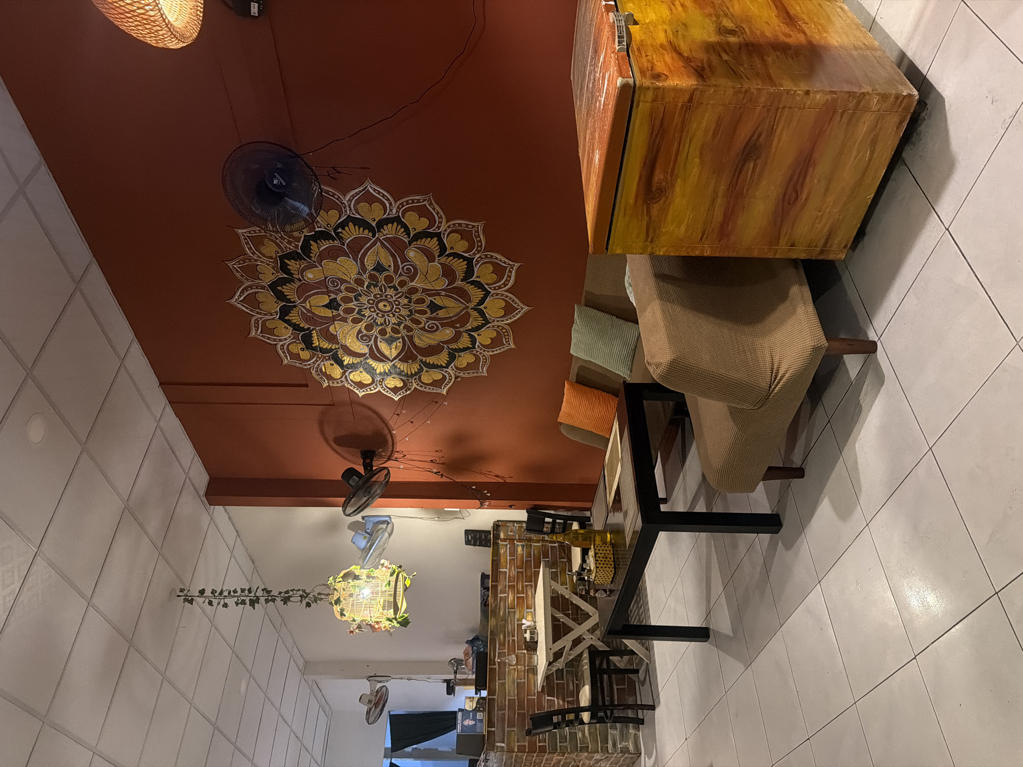 The image depicts the interior of a restaurant located in Bac Nha Trang Ward, Vietnam. The restaurant features a vibrant orange wall adorned with a large, intricate mandala mural. To the left of the mural, a black oscillating fan is mounted on the wall. In the foreground, a brown couch with an orange cushion sits next to a wooden chest painted in a faux wood grain pattern with orange and yellow tones.
Further back, a dark rectangular dining table is set with placemats and a bottle of water. Two black chairs are positioned at this table, and another table, smaller and square, with a white top and a wooden base, is situated to the left. This smaller table is accompanied by two black chairs. Behind these tables, a brick counter with items on top, including bottles and a possibly a cash register, is visible.
The ceiling is white and tiled. Hanging from the ceiling is a decorative cage-like light fixture adorned with artificial flowers and greenery. Two wall-mounted fans are visible on the left side of the image, one white and one blue. The floor is tiled with large, light-colored square tiles. The overall ambiance suggests a casual dining establishment with a decorative, possibly bohemian, theme.