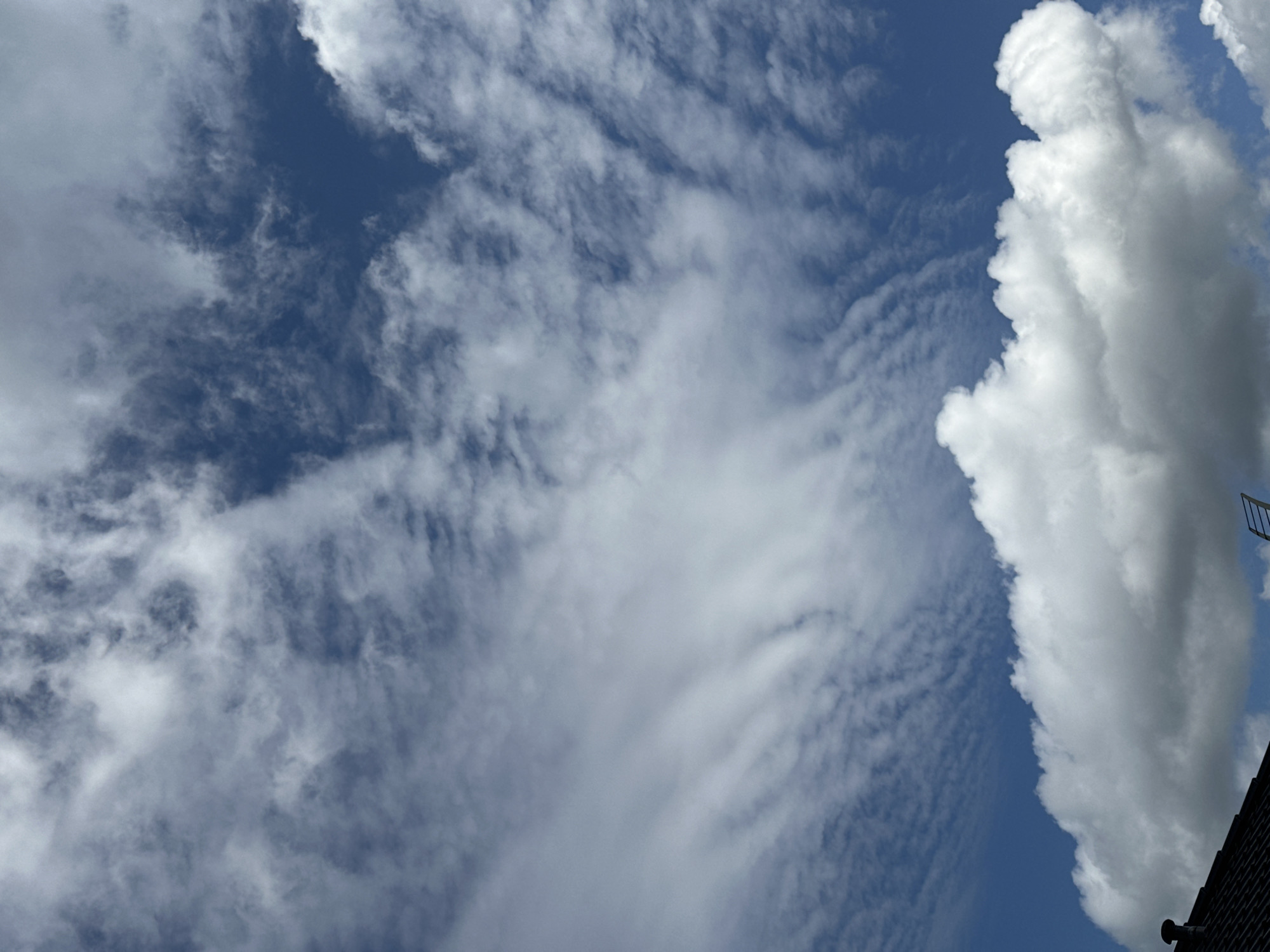 The image shows a bright blue sky with a variety of clouds. In the foreground, a large cumulus cloud billows across the lower third of the frame, with its base casting a shadow on what appears to be a dark roofline in the bottom left corner. The roof has some dark shingles and a small black chimney or vent. Above this, the sky transitions to wispy cirrus clouds that streak horizontally, creating a layered effect. These thinner clouds are more translucent, allowing the deep blue of the sky to show through. The overall impression is of a clear, sunny day with diverse cloud formations. The location is the city of Wisła, Poland.