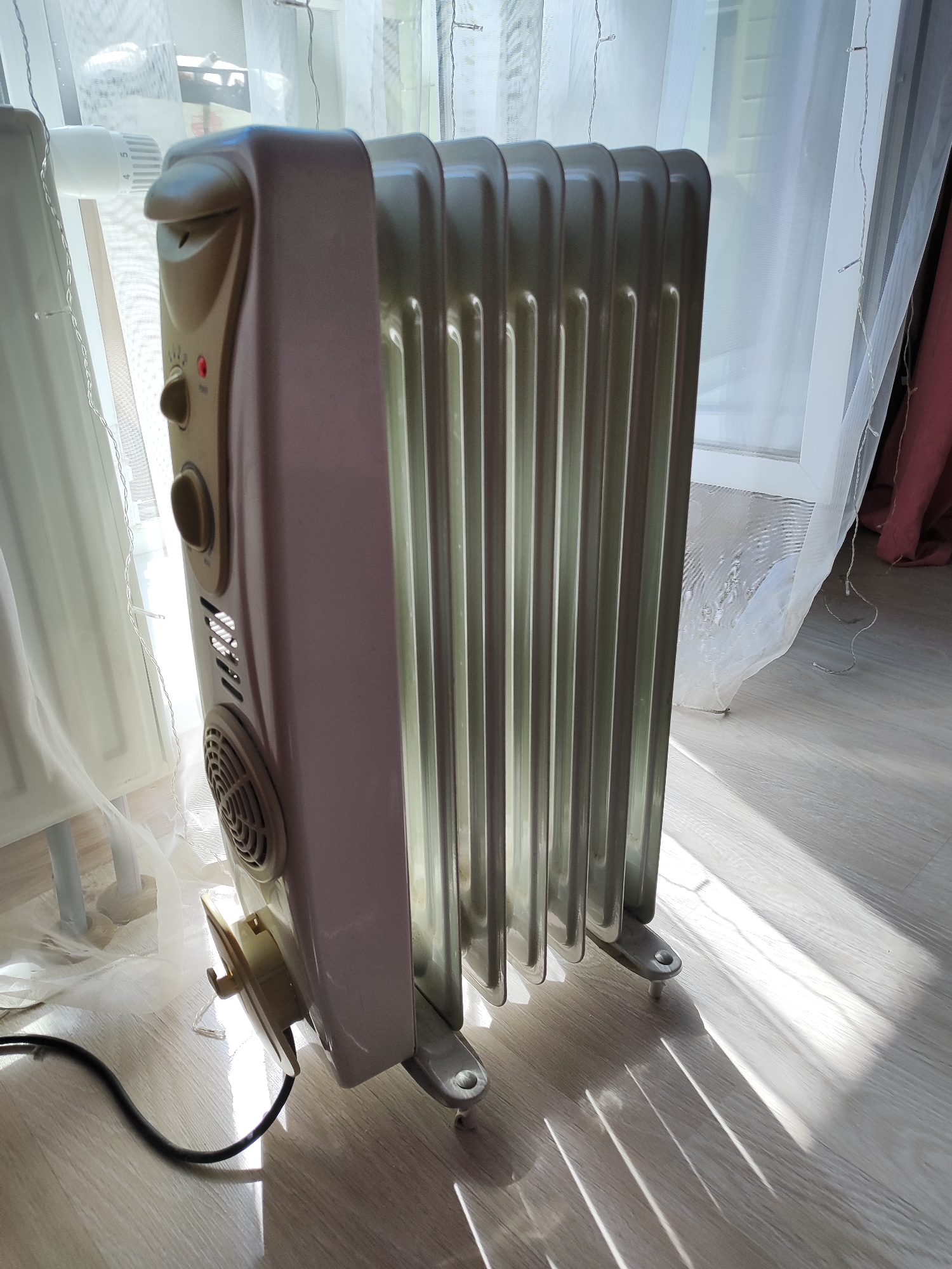 The image displays a close-up view of a white electric oil-filled radiator standing on a light-colored wooden floor. The radiator is positioned to the right of the frame, angled towards the viewer, with its fins clearly visible. The top of the radiator has a control panel with two knobs and a red indicator light, suggesting it is operational. Sunlight streams in from the left, casting distinct shadows across the floor and illuminating the radiator. Behind the radiator, sheer white curtains are partially drawn, and a hint of a window frame is visible. To the left of the radiator, another section of a white radiator is partially visible, along with what appears to be a thermostat. The overall scene suggests an indoor environment, likely a room in a residential building. There are no people or animals in the image. The lighting conditions are bright due to the sunlight. The scene appears to be in an apartment in Snezhinsk, Russia, as no other location information is available.