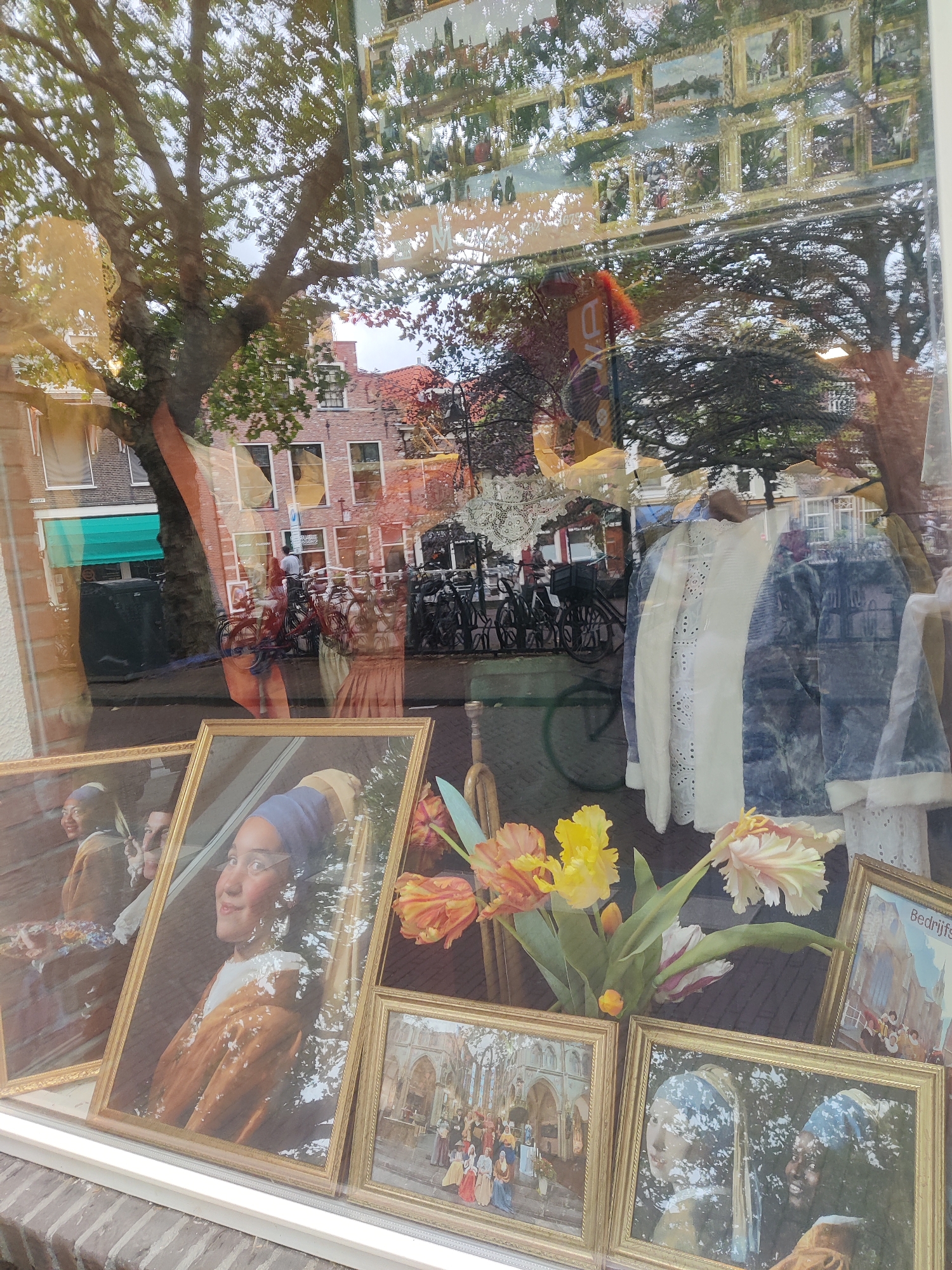 The image shows a shop window in Delft, Netherlands, with reflections of trees and buildings. In the foreground, there are several framed paintings. One painting depicts a woman wearing a blue headscarf and a brown garment, likely a reproduction of Vermeer's "Girl with a Pearl Earring." Another painting shows a group of people in historical dress gathered in front of a building, possibly a church. To the right, a framed picture with Dutch text, possibly "Bedrijfsfeest," and an image of a building is visible. Behind the paintings, a bouquet of tulips is arranged. The shop window displays clothing items, including a white broderie anglaise jacket and a blue patterned jacket. The reflections in the glass show a tree with green foliage, brick buildings, and bicycles. The overall impression is of a shop in a historic Dutch city, selling art and possibly clothing.