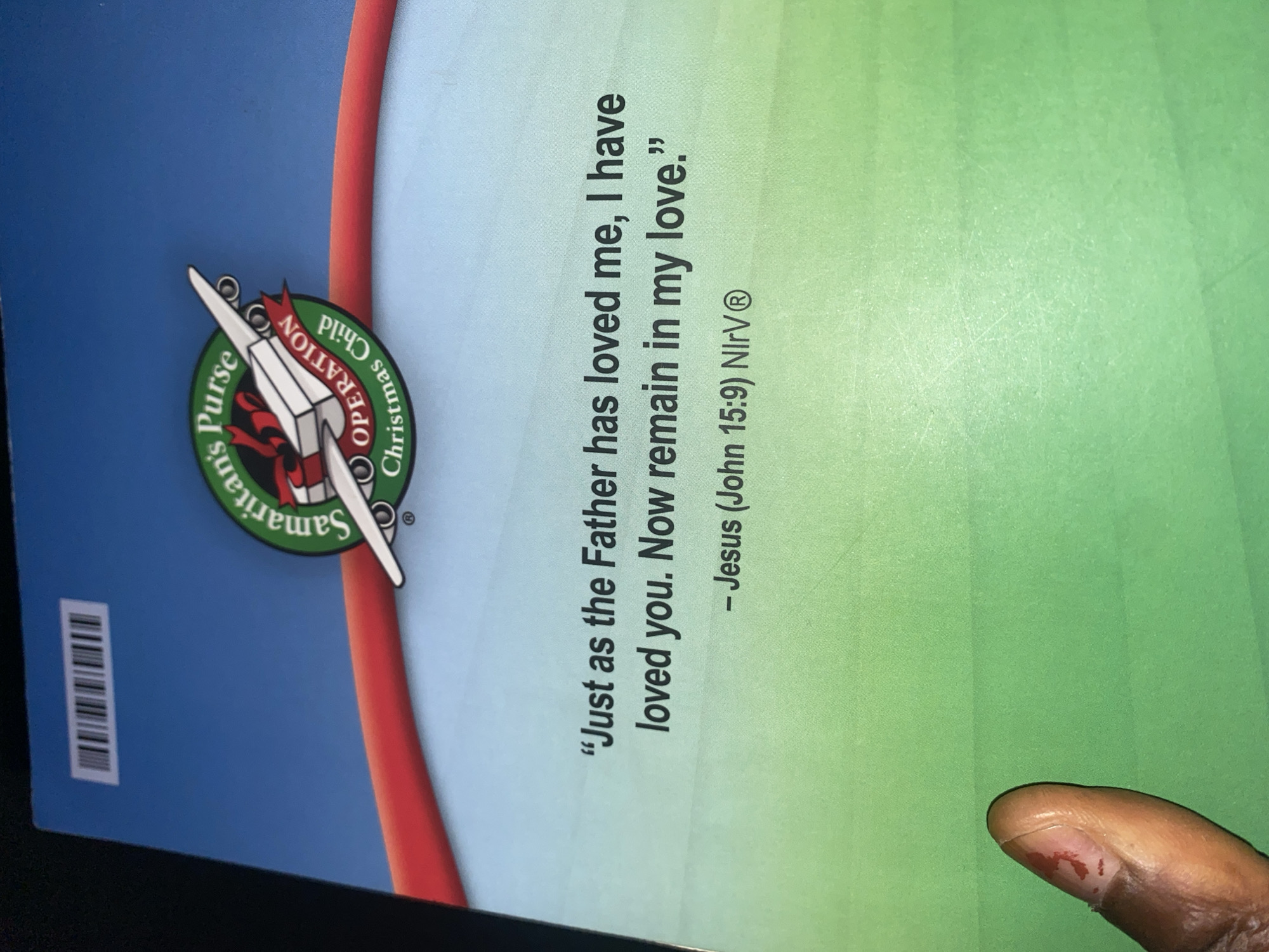 The image is a close-up, rotated view of a page with text and a logo. A person's finger with a visible bloodied cut is in the upper left corner, against a green gradient background. The text, oriented vertically, reads: "Just as the Father has loved me, I have loved you. Now remain in my love." followed by "- Jesus (John 15:9) NIV®" on the next line. To the right, a circular logo for "Samaritan's Purse Operation Christmas Child" is visible. The logo features a winged airplane and the organization's name. A barcode is present on the right edge of the page. The background transitions from green to light blue. The image does not contain information about Jauro Yinu, Nigeria.