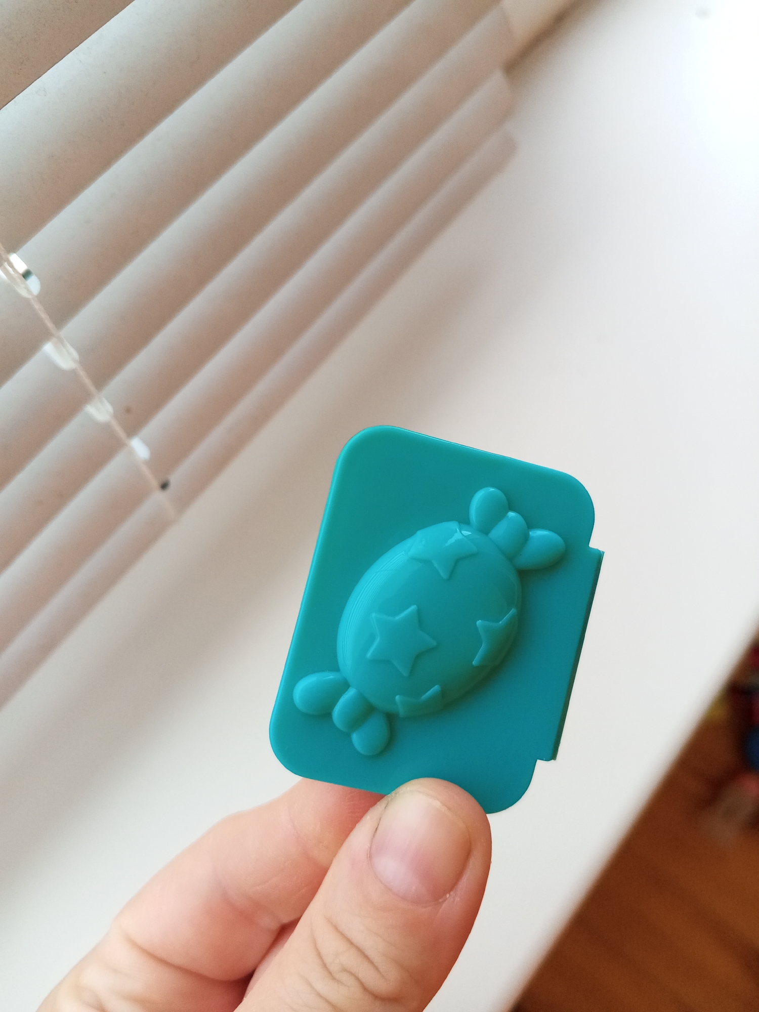 In this close-up image, a person's hand holds a teal-colored object with a playful design. The object is a flat, rectangular clip with a raised, cartoonish candy embellishment featuring three stars and two small, rounded projections resembling bunny ears. The clip itself has rounded corners, and a small notch is visible on its right edge, suggesting it's designed to attach to something.
The background is softly blurred, dominated by the white slats of window blinds, which are angled diagonally. A thin string, likely part of the blinds' mechanism, with clear beads, is visible to the left. The lighting suggests a bright, well-lit environment, possibly daytime, with the light casting a subtle shadow on the object and hand.
The image focuses on the tactile and visual aspects of the clip. The hand, with its visible fingernails and skin texture, is positioned to showcase the clip. The overall tone is casual and possibly related to everyday use or a decorative item. There is no visible text in the image. The provided location context of Chistopol, Russia, does not offer specific visual cues within the frame itself.