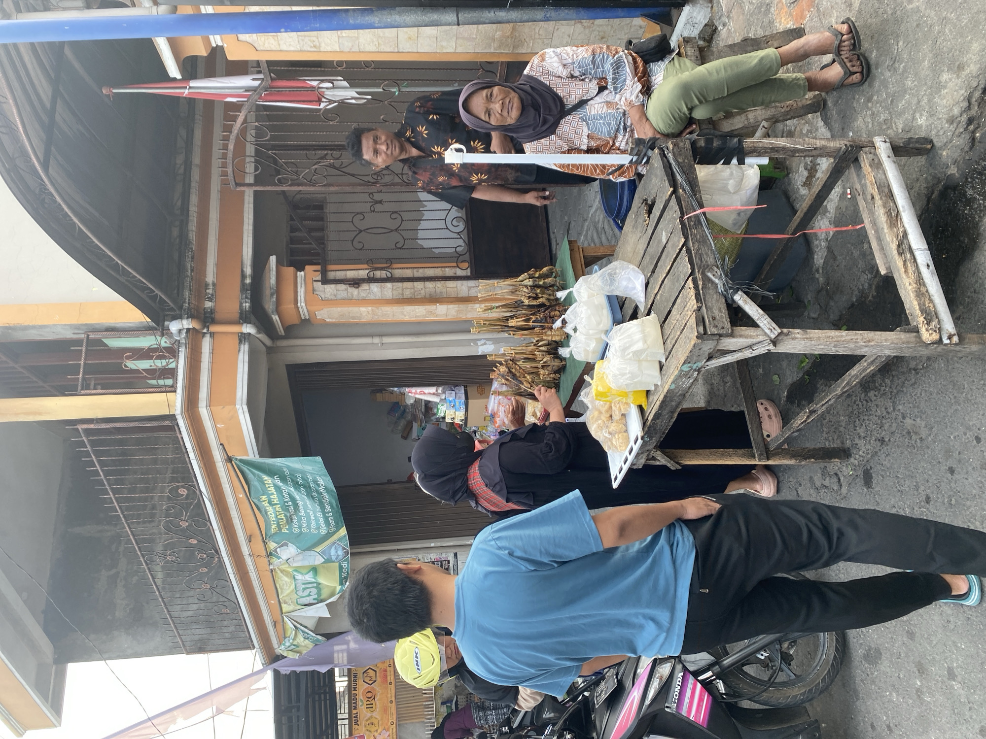 In the city of Kediri, Indonesia, a street scene unfolds with several individuals engaged around a small food stall. The stall features a rustic wooden table laden with food items, including skewers of cooked poultry and bags of what appear to be snacks. A woman in a hijab and a patterned shirt sits on a stool at the stall, facing the camera. Beside her, a man in a dark patterned shirt stands, looking towards the viewer. Another woman, also in a hijab, is partially visible behind the table, interacting with the food. In the foreground, a person wearing a blue t-shirt and black pants stands with their back to the camera, facing the stall. A motorcycle with a yellow helmet rests to the left. The background shows a building with orange and cream accents and a balcony with intricate metalwork. An Indonesian flag is visible near the entrance of the building. The ground is paved with asphalt.