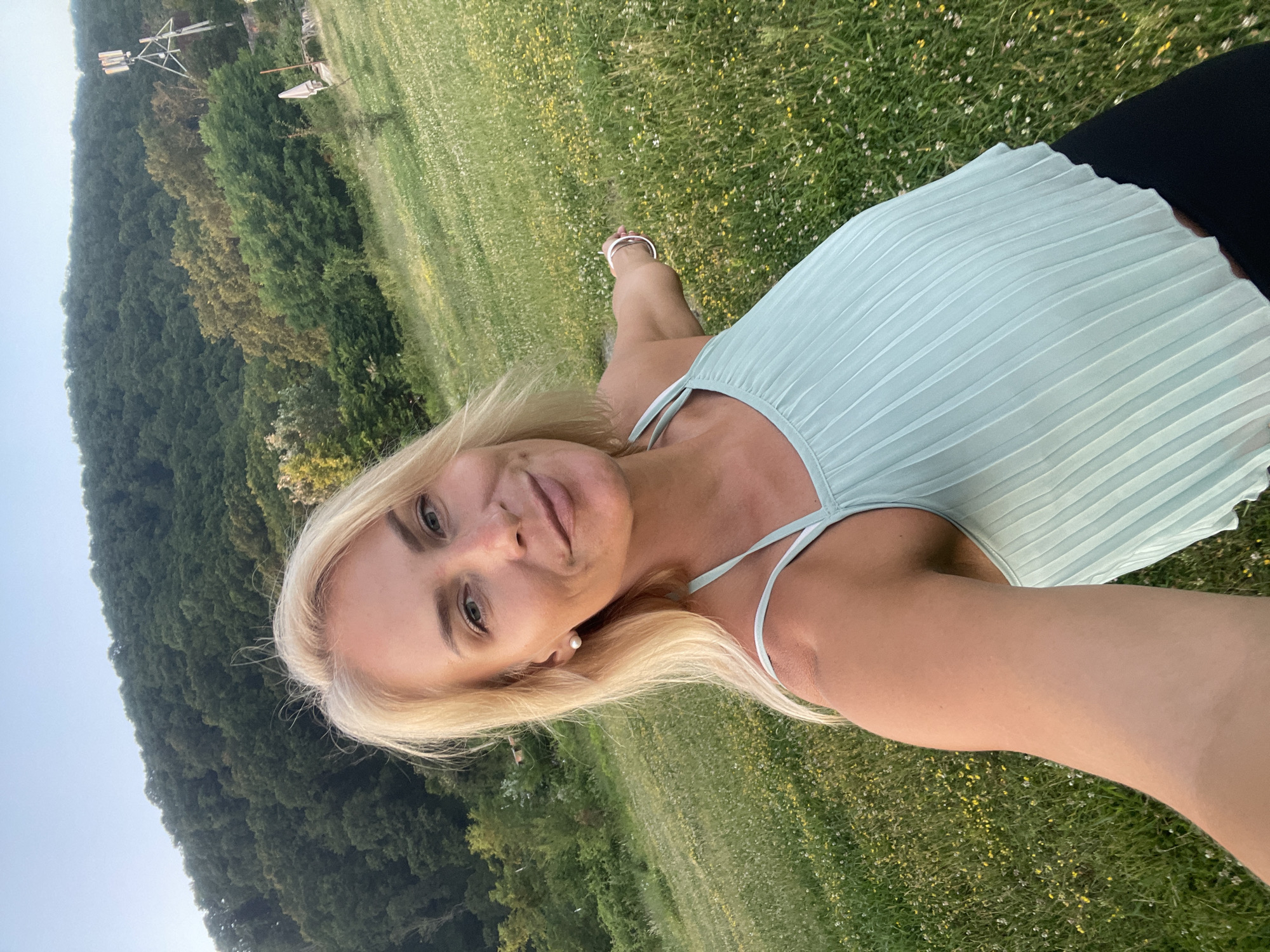The media file is a selfie of a blonde woman taken outdoors in a grassy field dotted with small yellow and white flowers. She is wearing a light blue pleated top with thin straps and black bottoms. Her right arm is extended, holding the camera. The woman has fair skin, blue eyes, and is wearing a pearl earring. She is smiling slightly, looking at the camera.
Behind her, the scene transitions into a dense forest covering a hillside. In the distance, on top of the hill, a metal structure with some lights is visible, possibly a radio tower or a weather station. The sky is a pale blue, suggesting it is either early morning or late afternoon. The overall atmosphere is serene and natural. The image is oriented vertically, suggesting it was taken in portrait mode. No text is visible in the image.