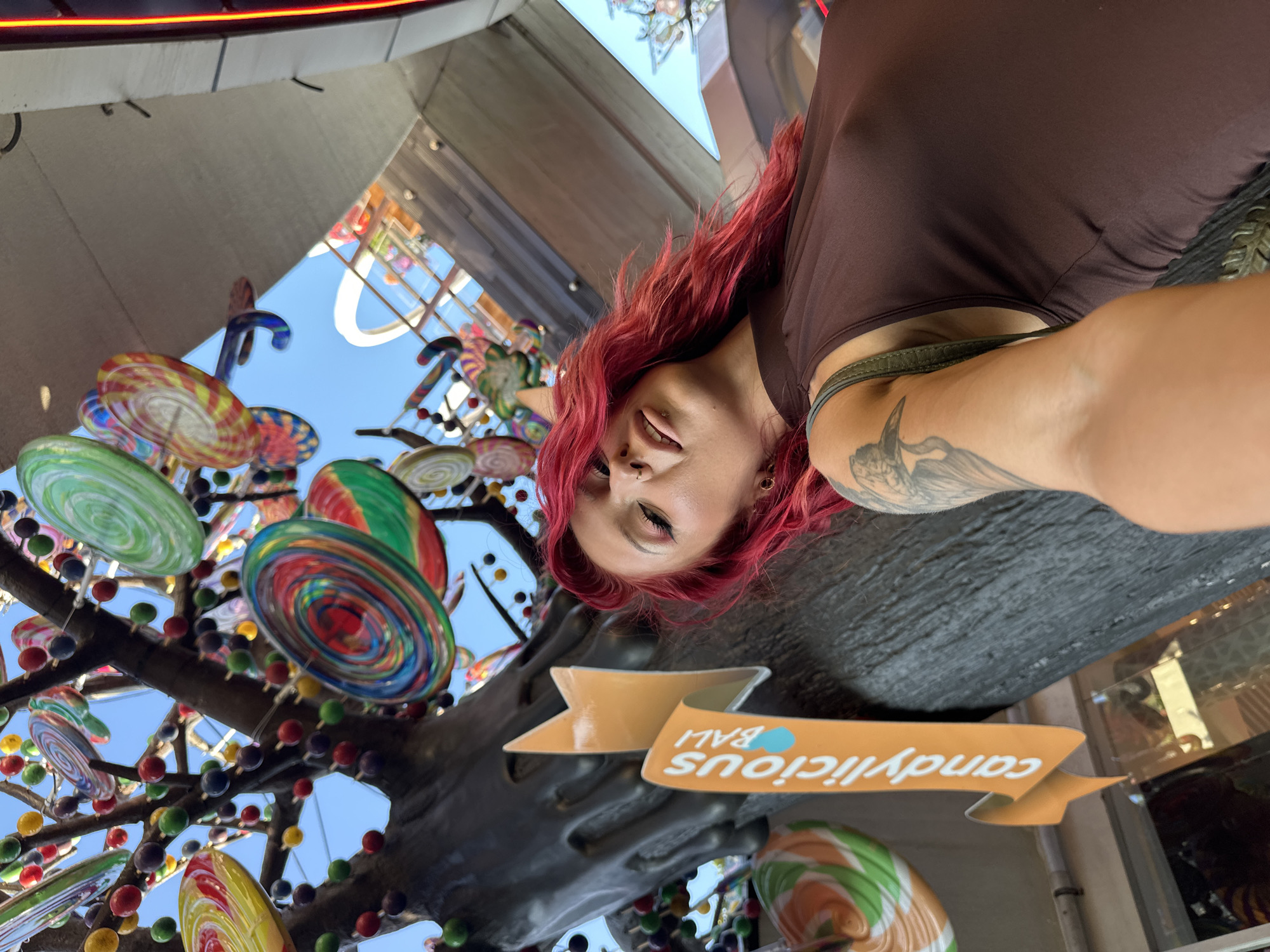 A woman with red hair, a brown tank top, and tattoos poses for a selfie in Kuta, Indonesia. Behind her is a "Candylicious Bali" sign. A large lollipop tree made of colorful lollipops, beads, and faux tree branches is also visible, set against a partly cloudy blue sky and a nearby building.