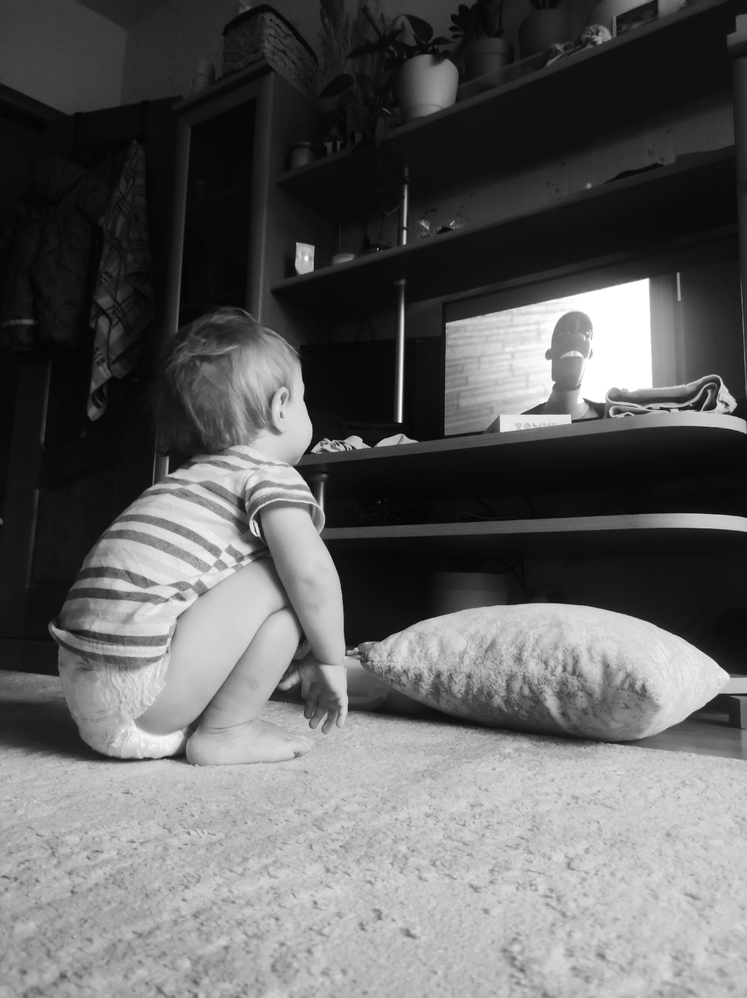 A black and white photograph depicts a young child sitting on the floor, facing a television screen. The child wears a striped shirt and a diaper. The television is on a shelving unit that contains plants and other objects. The background includes a cabinet. A pillow rests on the floor in front of the television. The city of Chaikovsky, Russia, is not visible in the photograph, so the location cannot be verified.
