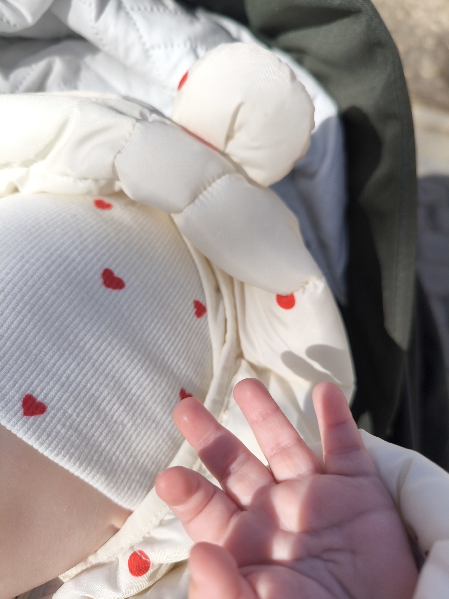 The image presents a close-up, high-angle shot centered on the torso and hand of a baby or young child, likely outdoors in Fryazino, Russia. The child is wearing a light-colored, possibly cream or white, ribbed top or bodysuit patterned with numerous small, red heart shapes. Over this, a puffy, cream-colored quilted jacket or blanket is visible, partially covering the child's body and arms.
The focus is drawn to the child's small, soft hand, which is slightly splayed with the palm facing upwards, extending towards the viewer. The fingers are short and plump, typical of an infant. Bright, natural sunlight illuminates the scene, casting strong highlights on the fabrics and the child's skin, and creating distinct shadows, notably across parts of the hand and the quilted material. In the background, a dark green or dark-colored fabric is visible, possibly part of a stroller, car seat, or another piece of outdoor equipment, suggesting an outdoor setting on a sunny day. The overall impression is one of innocence, warmth, and tenderness. There is no visible text in the image.