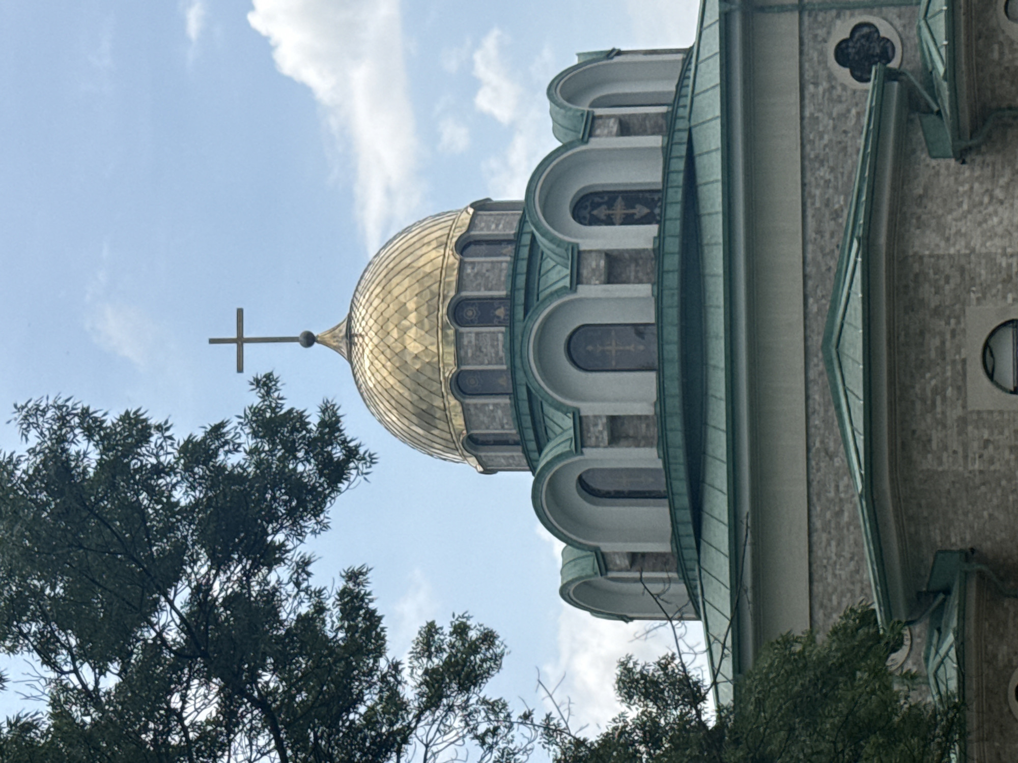 The image shows the exterior of a church with a golden dome topped with a cross. The dome is surrounded by smaller, arched structures with green roofs. The church walls appear to be made of light-colored stone blocks. Green foliage from a nearby tree partially obscures the upper left portion of the image. The sky is blue with scattered white clouds. The building is likely an Eastern Orthodox church due to its architectural style and the presence of a cross on the dome. Based on the architectural style, this is likely the St. Anastasia Monastery church in Burgas, Bulgaria. There are no people visible in the image.