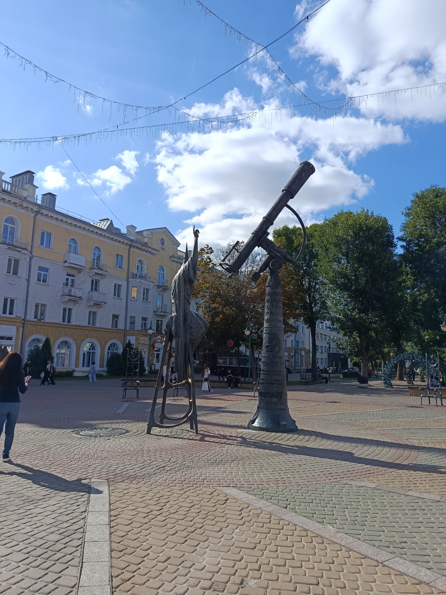 An eye-level, medium shot captures an outdoor public plaza in the city of Mahilyow, Belarus, on a clear, sunny day with scattered white clouds. The foreground is dominated by a cobblestone pavement patterned with light brown, gray, and reddish-pink rectangular stones, laid in concentric arcs radiating from the center of the frame. A long, narrow channel filled with rectangular stones runs diagonally across the lower center of the image, casting a distinct shadow.
In the mid-ground, a large, dark metal sculpture of a telescope on a cylindrical pedestal is the primary focal point. The telescope is angled upwards towards the sky, and the pedestal is adorned with what appear to be inscriptions in Cyrillic. To the left of the telescope sculpture, a smaller, metallic human-like figure stands on a multi-legged base, also with its arm raised. The paved area extends further back, with scattered benches, lampposts, and more people walking.
In the background, a multi-story building with a pale yellow facade and white trim, featuring arched windows and balconies, lines the left side of the plaza. Adjacent to the building are several evergreen trees. On the right side of the plaza, tall deciduous trees with green foliage create a backdrop. Further down the street, more buildings and trees are visible, and a few more people are seen walking or standing. A string of decorative lights is strung across the sky from the top left to the top right of the frame. One person, a woman with long dark hair, is walking away from the camera towards the left side of the frame, wearing a dark top and blue jeans. Other individuals are scattered throughout the plaza, engaged in passive activities such as walking or sitting. The overall atmosphere is one of a calm, sunny day in a well-maintained urban public space.