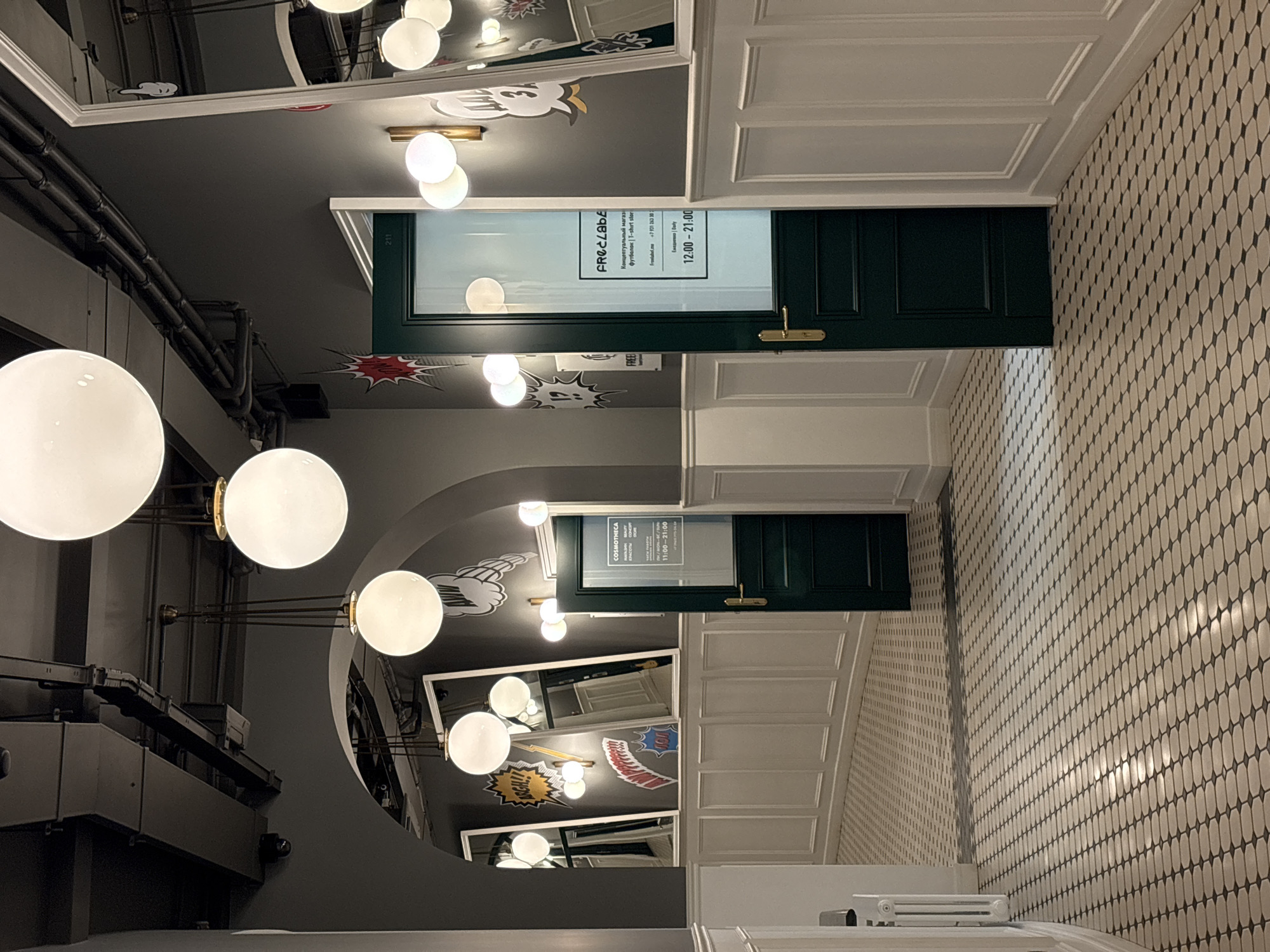 The image is a slightly distorted upward shot in an interior space. The walls have small oval tiling, a white trim along the ceiling, and green doors with glass windows and gold handles, each with black lettering, either "FRECLABE" or "COSMOTHECA", and schedules written in Russian. The ceiling is grey and has comic book-style text in speech bubbles, mirrors, and globe lights on brass fixtures. There are dark metal pipes running along the wall above the globe lights. Based on the text "ARGH!!", I can determine that this is likely somewhere on Ligovsky Prospekt in Saint Petersburg.