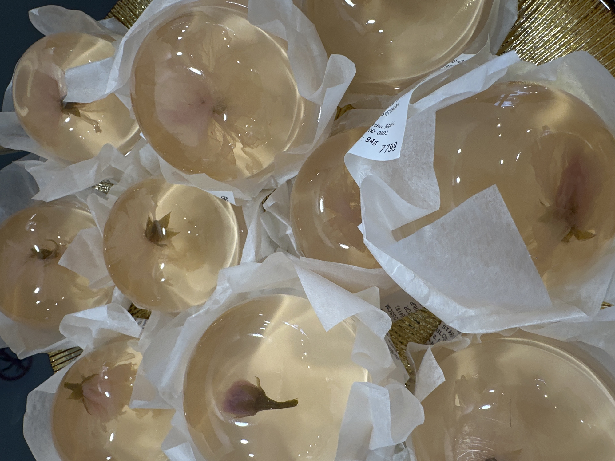 The image shows a collection of clear, dome-shaped candies or desserts, each individually wrapped in white tissue paper. Inside each dome is what appears to be a flower or bud, possibly an edible flower. These items are presented in a gold-colored, woven basket. A price tag is visible, displaying the price '7799' and the weight '84g', along with partial text that includes 'OSSO'. The origin is “KITCHOAN cho Kitaku 00-0803” which is a Kyoto, Japan address. The scene is indoors. There are no people visible in the image. The environment appears to be a shop or market setting, given the presentation and packaging of the items.