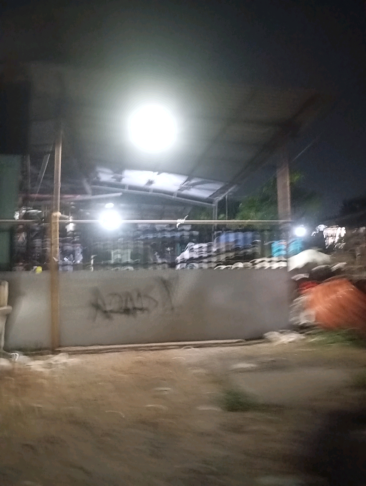 The image depicts an outdoor scene at night, illuminated by a bright overhead light. In the background, there appears to be a covered structure with corrugated roofing. Various objects are visible within and around this structure, including what look like stacked tires or similar items. A concrete wall with graffiti is centrally located in the foreground, obscuring some of the background details. The ground in front of the wall is composed of dirt and some scattered debris. To the right of the wall, an orange, possibly tarp-covered object is visible. The overall environment suggests a possibly industrial or storage area. The location is specified as Babelan, Indonesia.