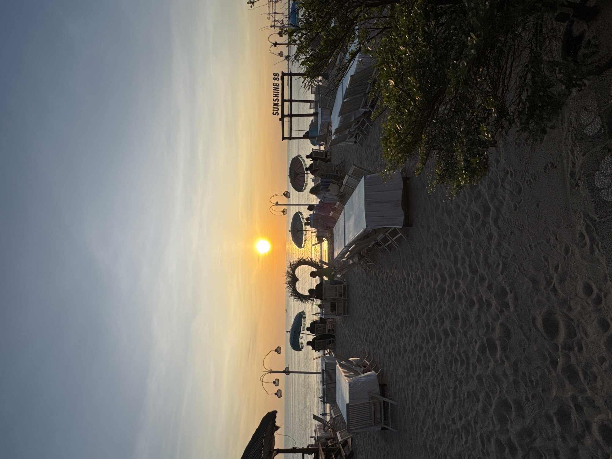 The image captures a sunset at Kuta Beach in Indonesia. A beachside scene features several tables and chairs set up on the sand, some covered with white tablecloths. People are seated at some of the tables, partially silhouetted against the setting sun. A decorative heart-shaped structure is positioned on the beach, with individuals seated nearby. Beach umbrellas provide shade over some tables. A sign reading "SUNSHINE 88" is visible in the background. The sky transitions from a light blue to an orange hue near the horizon where the sun is setting, creating a warm glow across the scene. The sand is textured with footprints and other marks. Trees are visible on the right side of the scene.