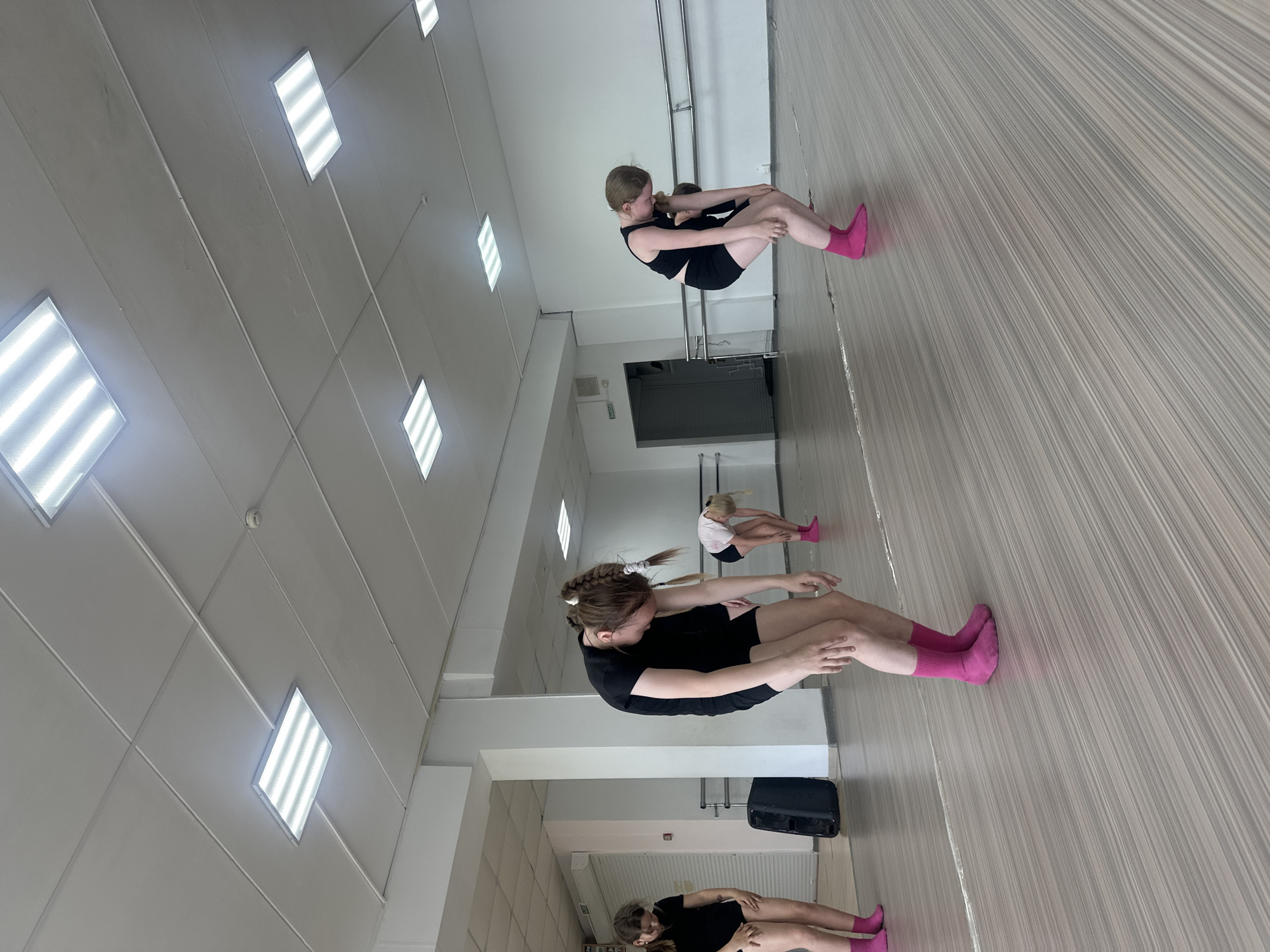In an indoor dance studio located in Votkinsk, Russia, three young girls are engaged in a stretching exercise. They are seated on the wooden floor, knees bent, and hugging their legs. All three girls are wearing black leotards and bright pink socks. The studio is well-lit with multiple fluorescent square lights in the white tiled ceiling. A metal ballet barre is affixed to the wall in the background. A speaker is situated near the center of the room.