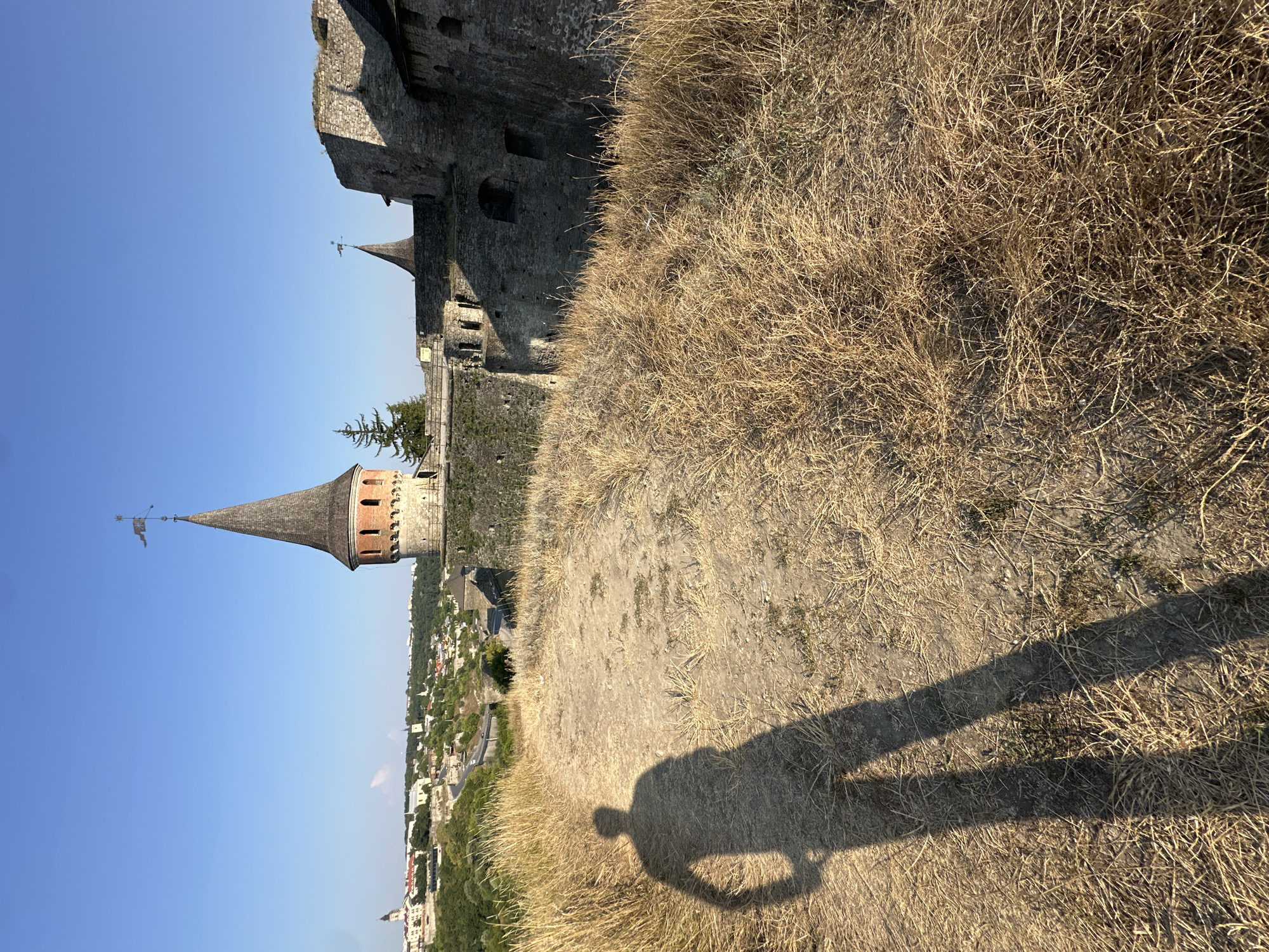 The image depicts a sunlit view of the Kamianets-Podilskyi Fortress in Ukraine. The foreground features dry, golden grass and a pathway. A long shadow of a person, likely the photographer, is cast on the path, suggesting it's a sunny day. In the background, the imposing stone walls and a conical tower of the fortress rise against a clear blue sky. Part of the city, with its buildings and greenery, is visible in the distance, nestled in a valley. The fortress appears well-preserved, with a weathered, ancient aesthetic. The overall scene conveys a sense of historical grandeur and natural beauty.