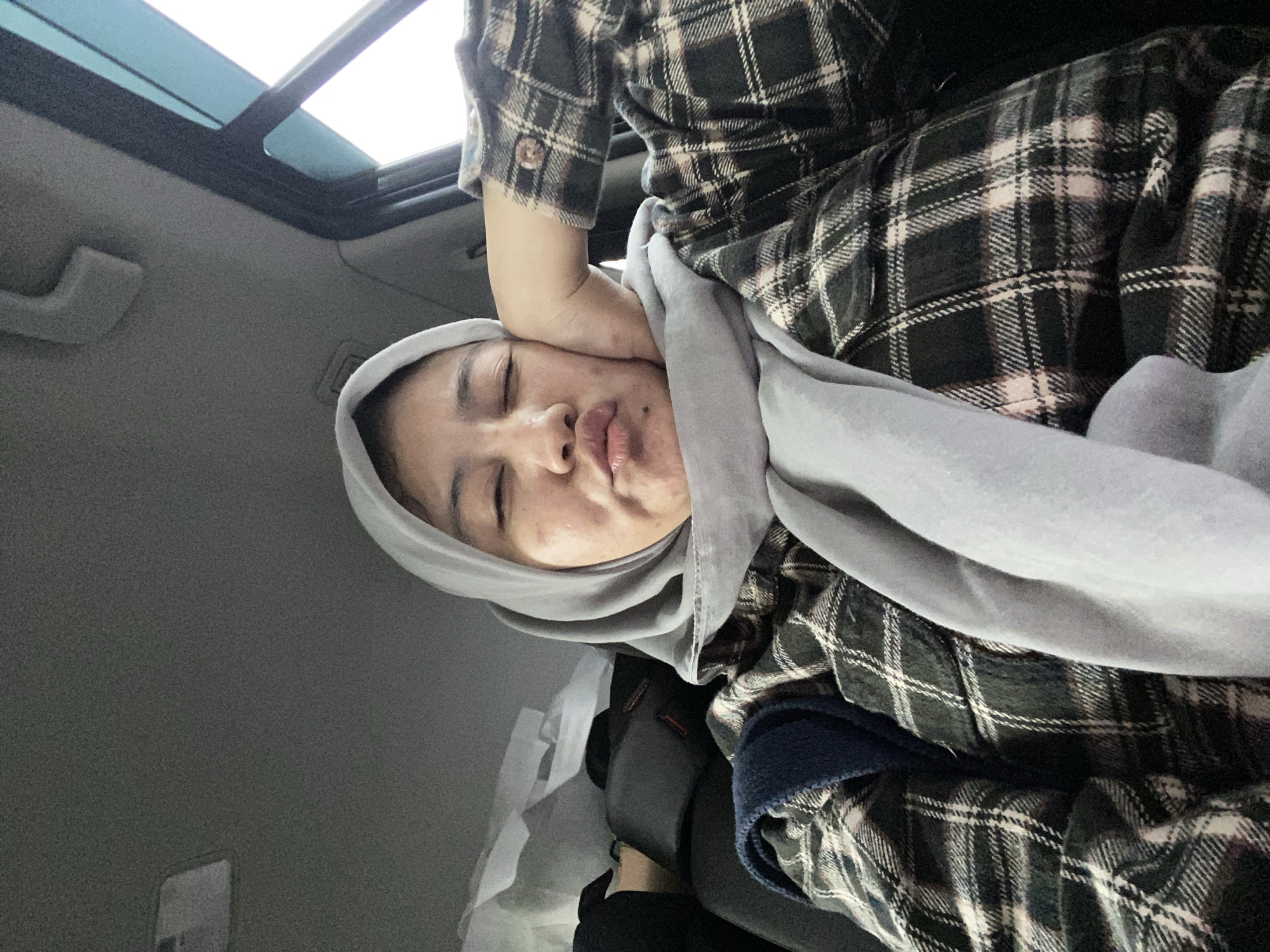 A medium shot captures a young woman with her eyes closed and lips puckered, resting her right cheek on her hand while seated in the passenger seat of a vehicle. She is wearing a light gray hijab and a dark plaid shirt with white and light pink lines. The background reveals the interior of a car, with glimpses of the ceiling, a window, and luggage or bags in the back. The lighting suggests an overcast day. The image appears to have been taken in Banjarbaru, Indonesia.