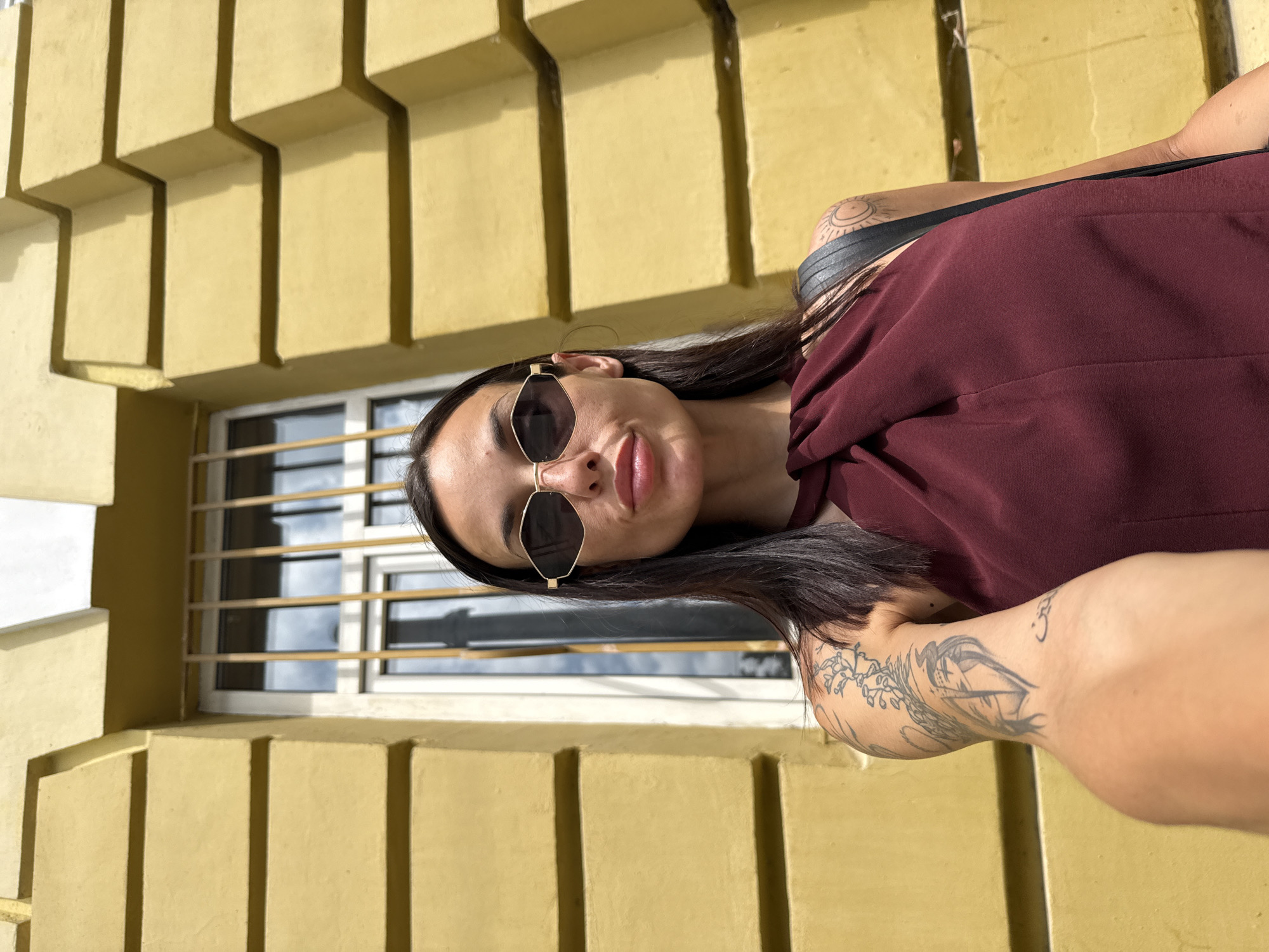 A young woman is captured in a close-up, portrait-style shot, leaning against or standing next to a bright yellow building. The image is oriented vertically, suggesting it was originally taken with a rotated camera. The building facade features prominent, geometric block-like protrusions that create strong, angular shadows, indicating bright sunlight. Behind the woman, a window with white frames and horizontal metal bars reflects the clear sky.
The woman has long, dark hair and is wearing distinctive gold-framed, dark-lensed hexagonal or cat-eye shaped sunglasses. She wears a dark reddish-brown or maroon top with a wide neckline. Several tattoos are visible: a circular, sun-like design with radiating lines on her left shoulder/upper arm, and a larger, intricate tattoo on her right forearm depicting a ship alongside flowering branches. A small "Om" symbol is also visible higher on her right arm. She has a confident and relaxed expression, with a subtle smile on her lips. The bright lighting suggests it is daytime, likely midday or early afternoon, on a clear, sunny day. The setting is an urban environment, specifically located in Pleven, Bulgaria. No discernible text is visible in the image.