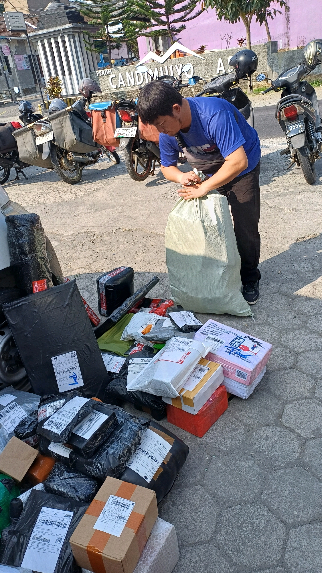 The image depicts a man in a blue t-shirt and black pants, wearing a fanny pack, in front of a building with the text "PERUM CANDIMULYO" in large letters. He is standing on a paved surface, surrounded by numerous packages and boxes of various sizes. Some packages are wrapped in black plastic, while others are in cardboard boxes and some are covered with white or light-colored plastic. A large, light-colored sack is in front of the man, and he appears to be holding onto it. Several motorcycles are parked behind him, some with cargo baskets. The environment suggests an outdoor, possibly commercial or residential area, with a sunny day and trees visible in the background. The overall scene suggests a delivery or logistics operation.