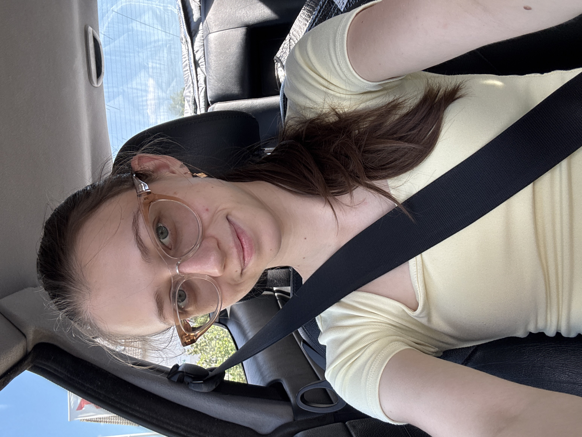 A young woman is sitting in the driver's seat of a car, wearing a light yellow top and large, translucent-framed glasses. She has her hair in a low ponytail and is smiling at the camera. A black seatbelt is visible across her chest. The interior of the car, including black leather seats and the car's headrest, is visible in the background. Outside the car, through the windows, a bright blue sky and some greenery can be seen. The scene appears to be taken during daylight, possibly on a street in Wörth am Main, Germany, given the overall context.