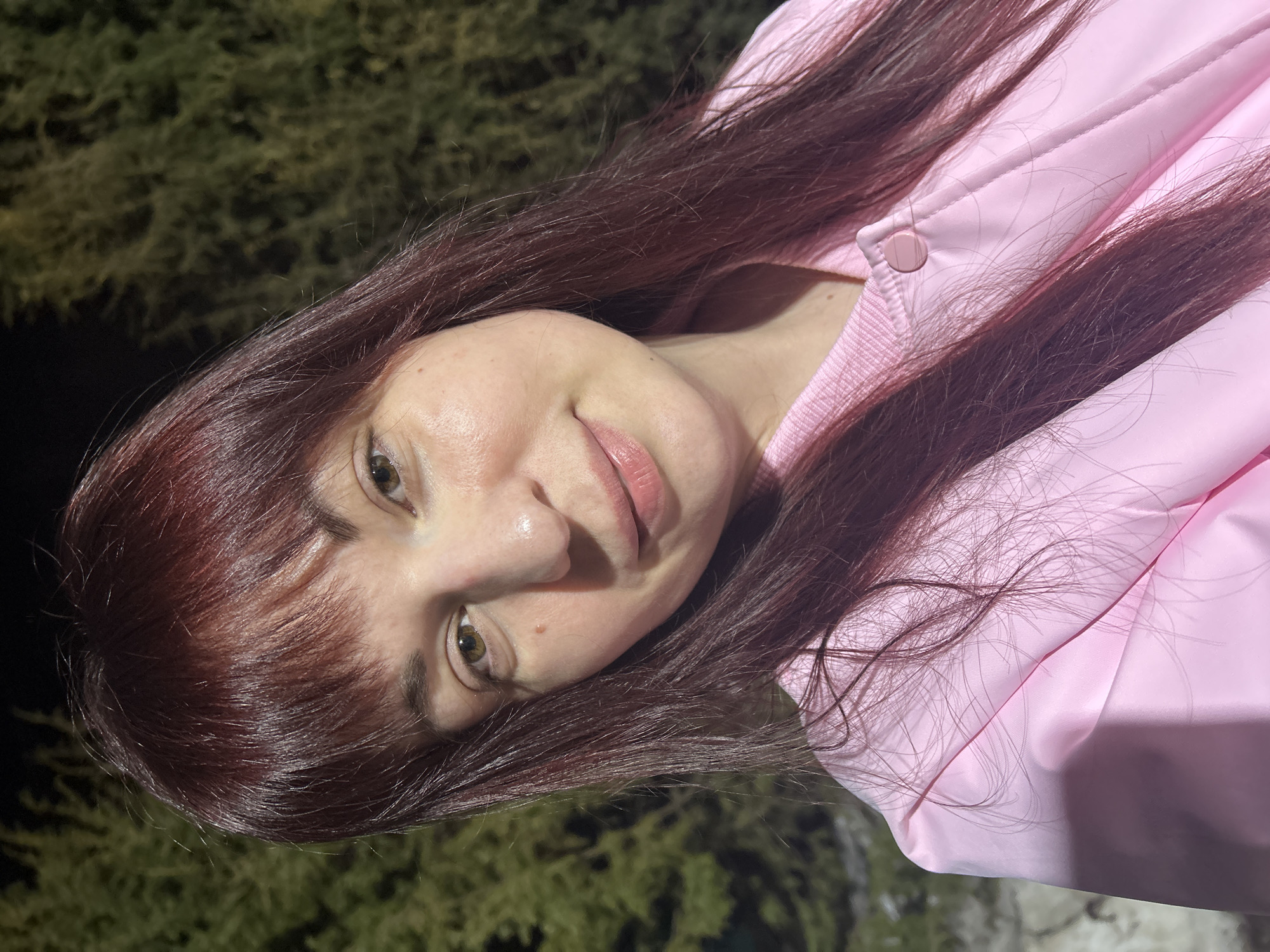 A woman is wearing a pink jacket. She has brown hair with bangs and green eyes. The background features some greenery and a dimly lit environment. It is not possible to determine the location in the city of Фоки, Russia from this image.