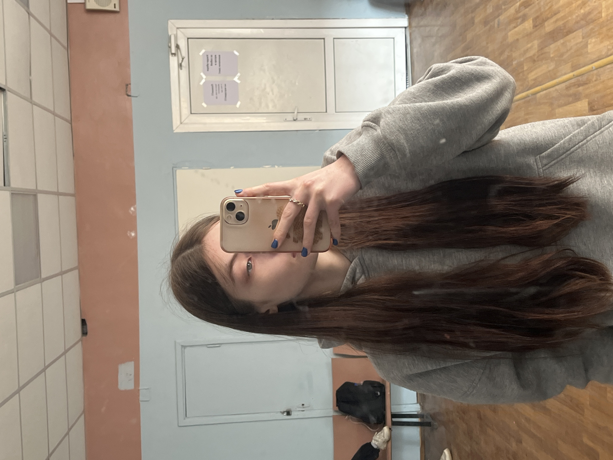 A person with long brown hair wearing a gray hooded sweatshirt is taking a selfie in a mirrored room. The room has white tile walls and a peach-colored vertical stripe. A white door with signage is visible. A wooden floor is present, and some items are on the floor near the mirror, including what appears to be a black bag and white shoes. Based on the architectural style and the interior details, the location may be in Saint Petersburg, Russia.