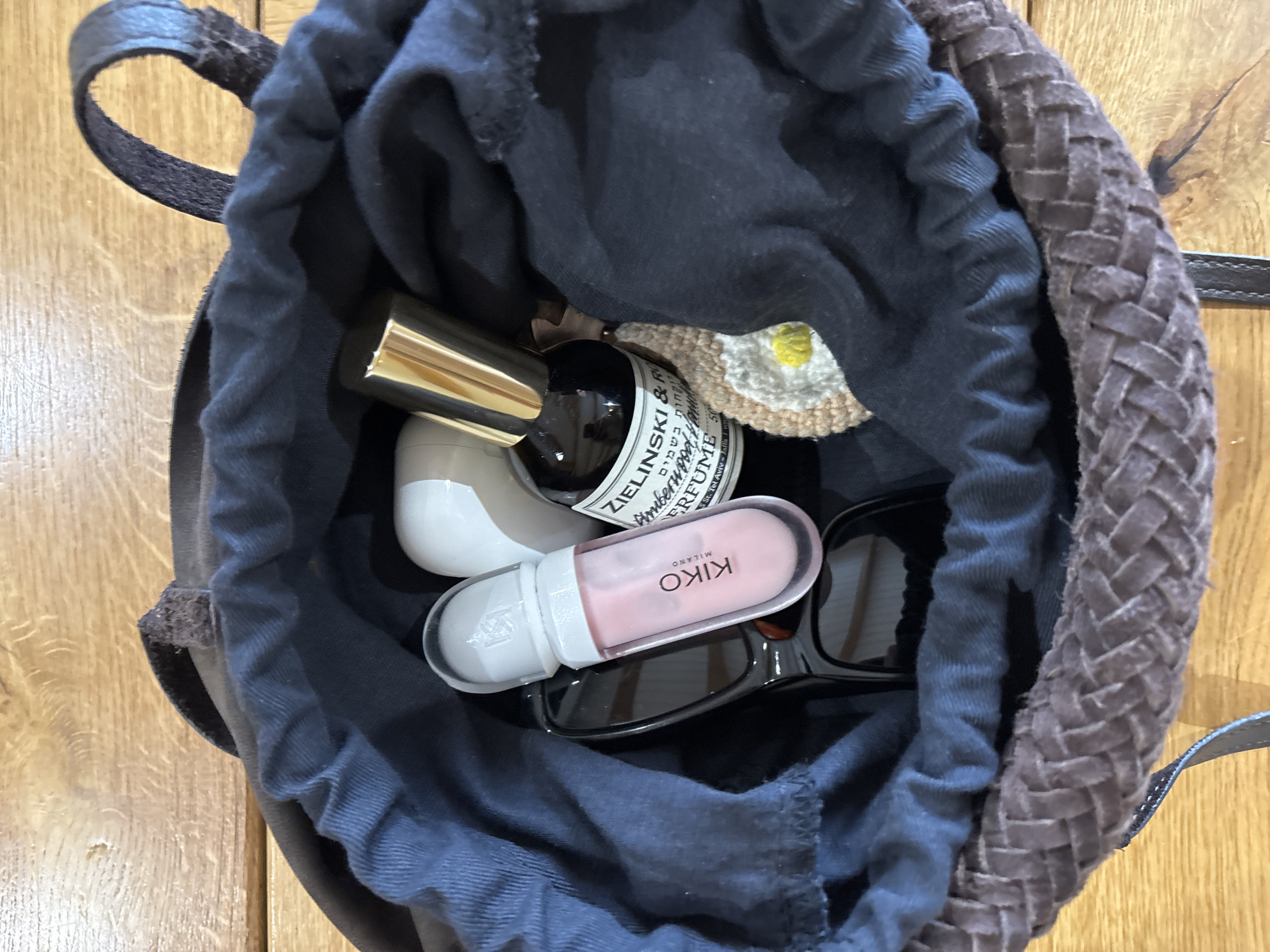 The image shows the open top of a dark blue drawstring bag. Inside the bag, several items are visible, including a bottle of Zielinski & Rozen perfume with a gold cap, a white and pink Kiko Milano lip gloss, a pair of sunglasses, a white object that appears to be a small container or electronic device, and a crocheted daisy-shaped object. The bag has a braided brown leather trim and handles. The background is a wooden surface. There is no indication of the location of Chaikovsky, Russia.