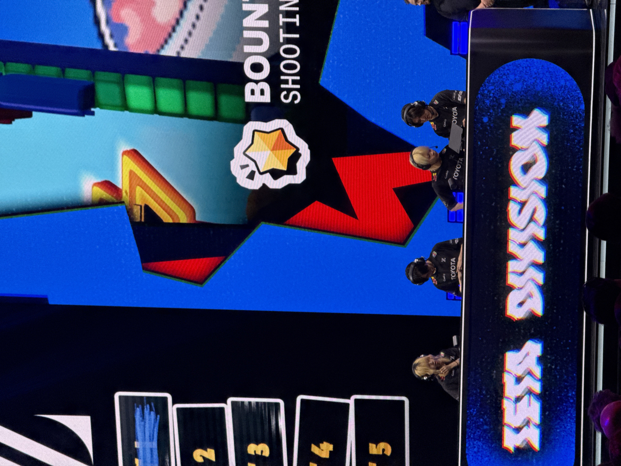 The image depicts a live broadcast of a gaming event, seemingly the "Brawl Stars" Championship, taking place in Berlin, Germany.
The main focus is the screen displaying game graphics with a "Bounty Shooting" mode, featuring elements like a star icon and colorful abstract shapes. The screen also presents numbered cards (2, 3, 4, 5) along the bottom edge.
On the right side of the screen, there is an overlaid graphic that reads "ESM DIVISION."
In front of the screen, players wearing headsets and branded shirts (Toyota) are actively engaged in the game, indicating a competitive tournament setting.
Audience members are also visible in the shot.
