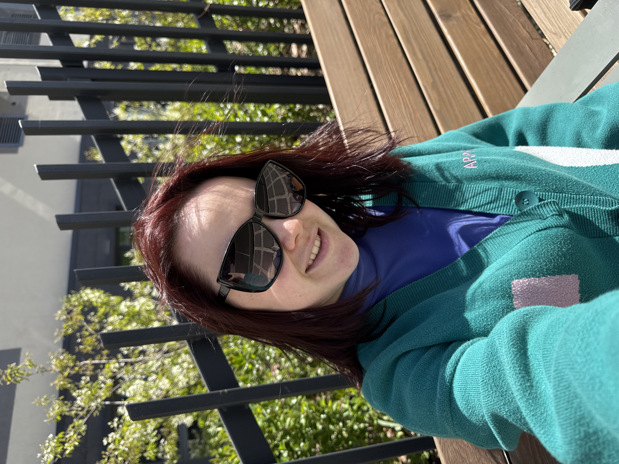 A woman with auburn hair and sunglasses is smiling at the camera. She is wearing a green cardigan over a blue turtleneck shirt. The cardigan has a white stripe across the chest and pink lettering that says "APP". She is sitting on a wooden bench with black metal railings behind her. The background includes greenery and a building. The scene is outdoors in daylight. The location in the city of Izhevsk, Russia, cannot be determined from the image.