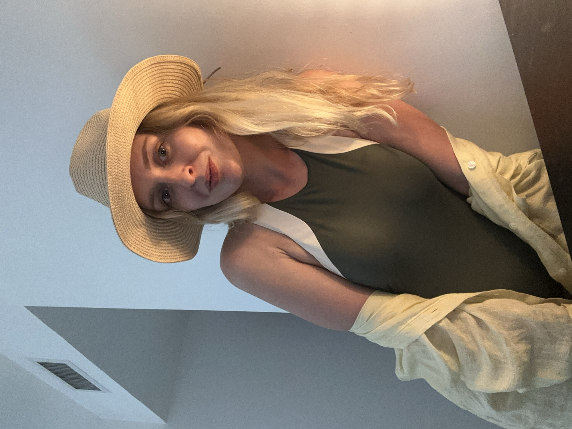 The image shows a woman wearing a tan straw hat, a dark green tank top with a white accent, and a light yellow button-down shirt. She is looking at the camera with a slight smile. The background consists of a white ceiling and a gray wall with an air vent. The location is Unknown, Unknown.
