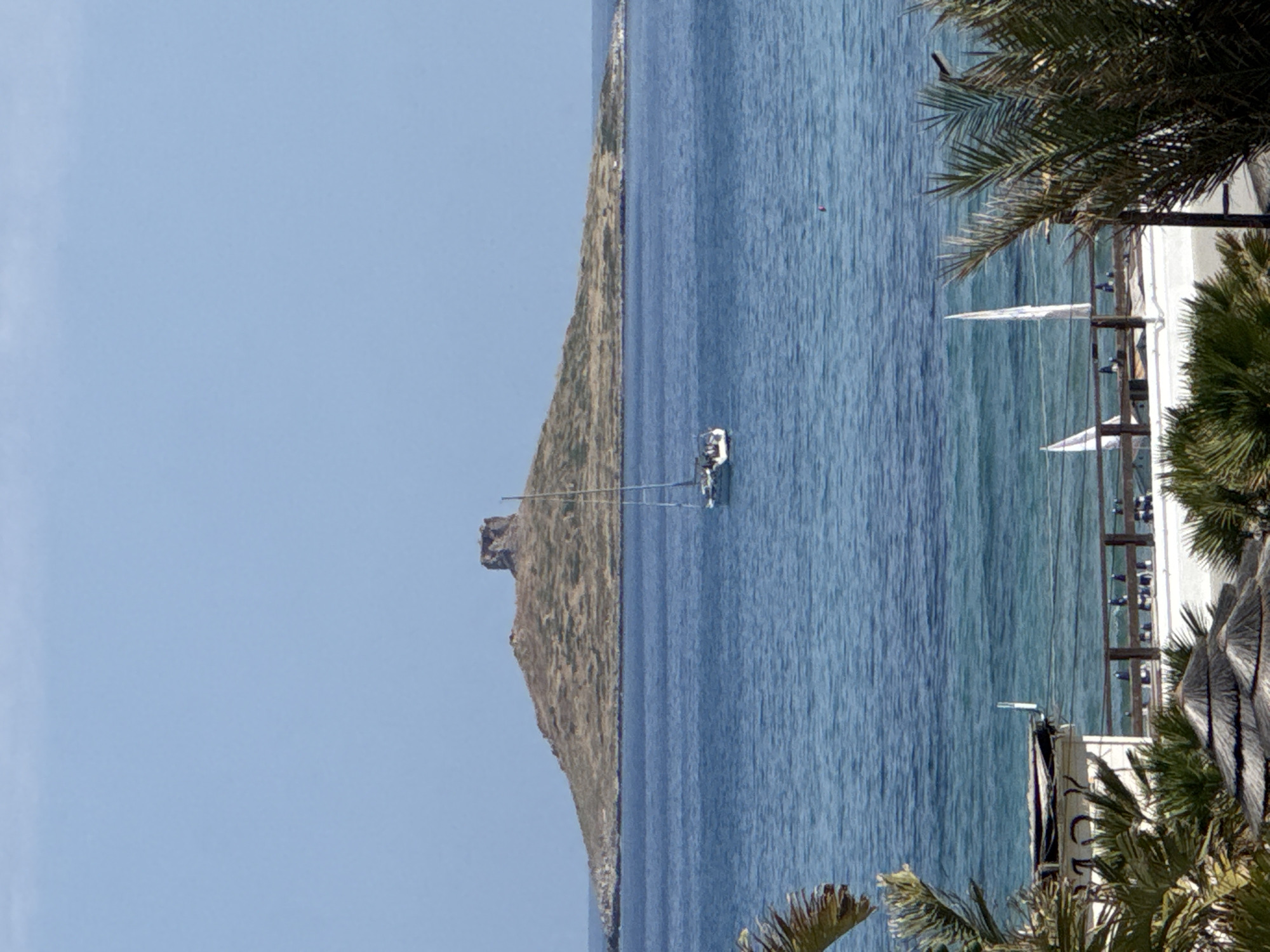 The image captures a peaceful coastal scene in Capaci, Italy, under a clear, bright blue sky. A calm, deep blue sea dominates the middle and right sections of the frame, exhibiting gentle ripples on its surface. To the left, a large, rugged landmass or island extends across the horizon, characterized by rocky formations and sparse, brownish-green vegetation. A distinct, dark rocky outcrop stands prominently near the center of this distant land.
On the water, a white sailboat with its mast upright is visible in the mid-distance. Closer to the right foreground, a beach area is framed by lush green palm trees whose fronds provide shade. Beneath these, several white beach umbrellas or awnings are set up, accompanied by what appear to be beach chairs and indistinct figures, suggesting people are relaxing. Parts of a wooden structure, possibly a pier or a beach club building, are also present. A small red buoy floats in the water near the beach.
The scene depicts daytime, likely morning or early afternoon, with abundant sunlight and fair weather, conveying a sense of leisure and tranquility. Partially visible white text on a foreground structure reads "CLU...", suggesting it is part of a sign for a beach establishment.