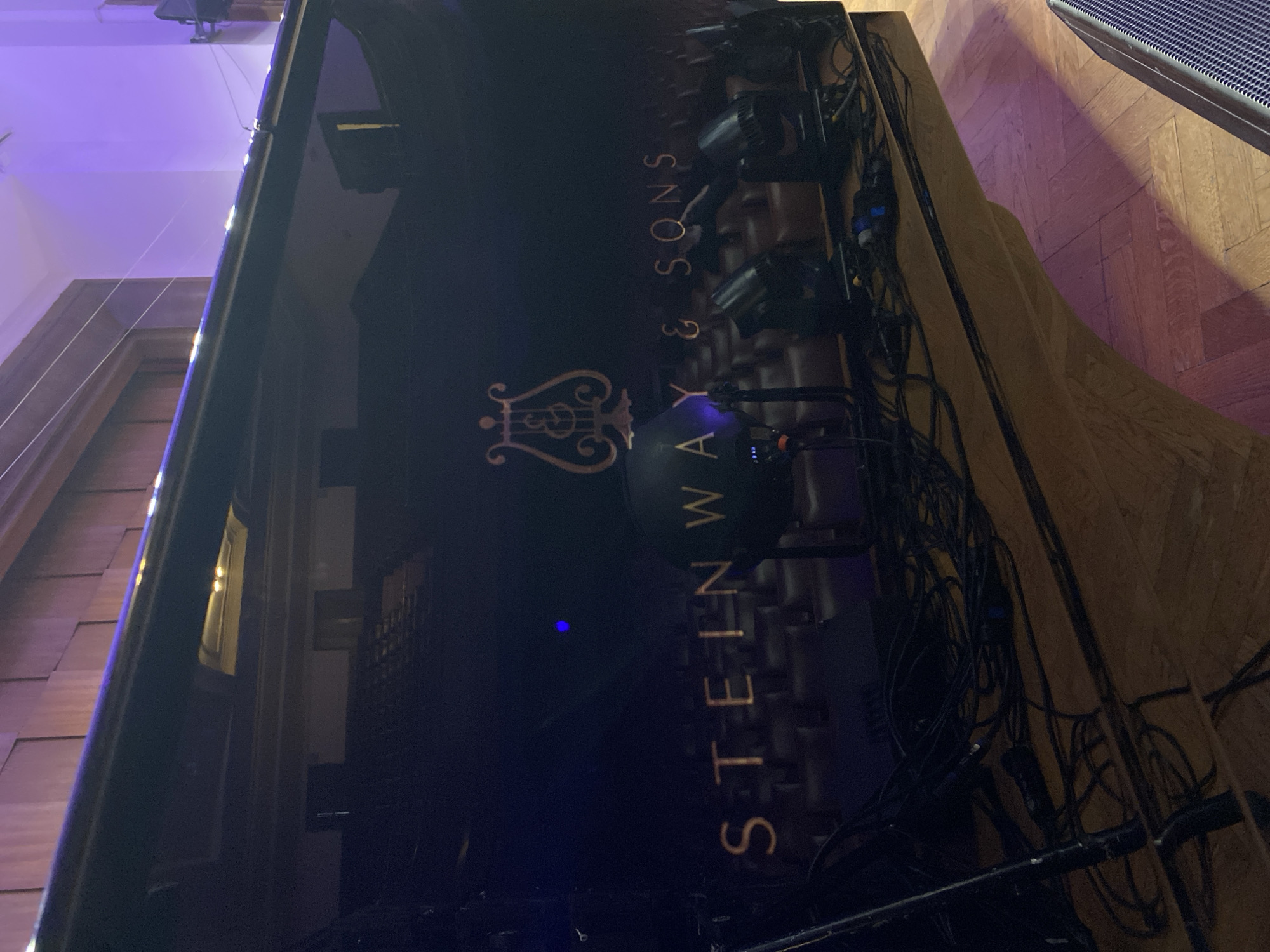 The image shows a partial view of a Steinway & Sons grand piano, likely on a stage setup. A rigging with stage lights is mounted behind the piano, with many black cables visible. The setting appears to be indoors, possibly a concert hall or auditorium. The floor has a parquet wood pattern. It is not possible to determine the precise location in Belgrade, Serbia, from the image alone.