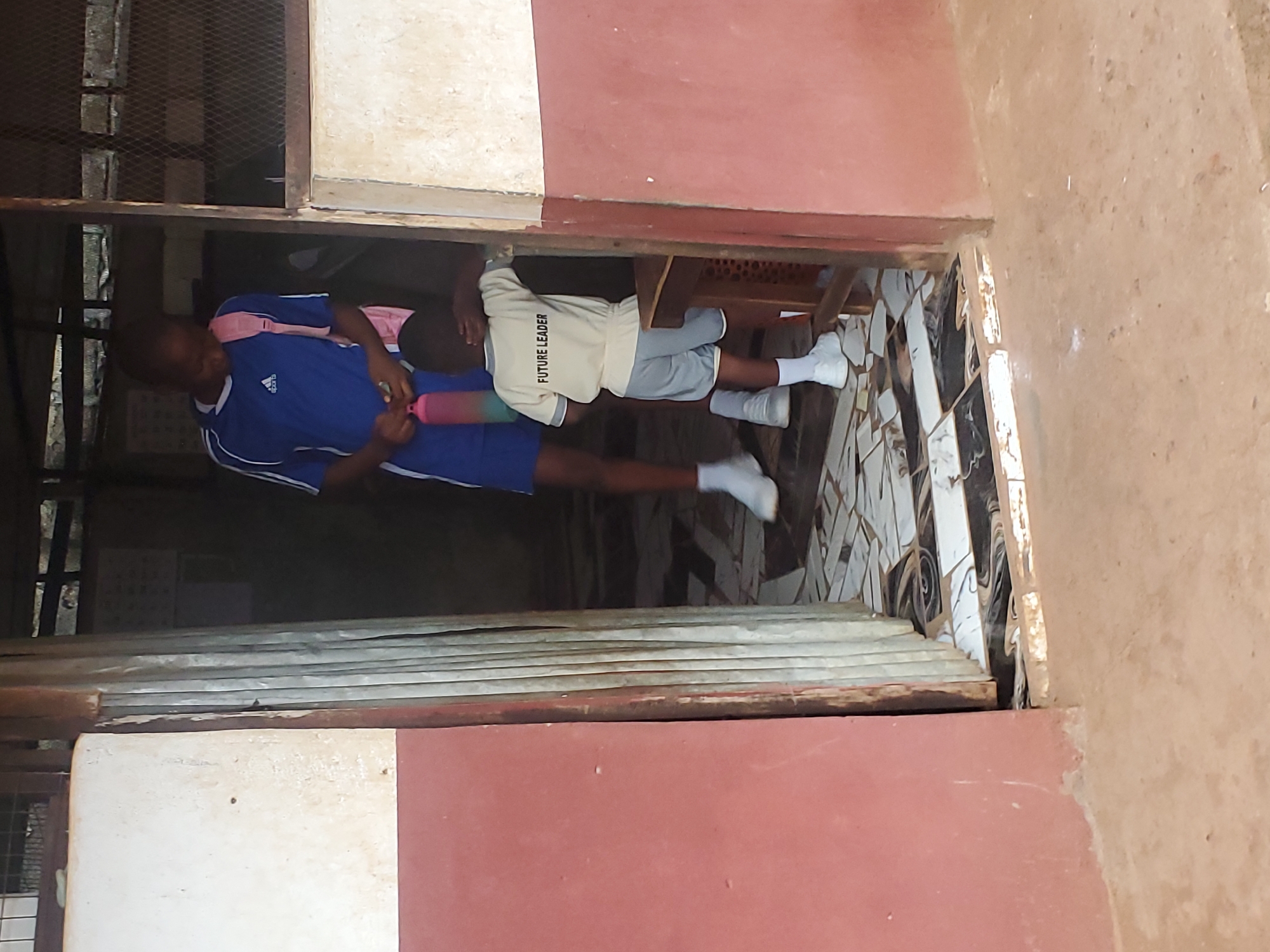 A photograph taken in what appears to be Ukwu-Abwa, Nigeria, shows two young individuals positioned within the confines of a building structure. The building features a reddish-brown and white exterior. One of the children is clad in a blue athletic shirt with white stripes on the sleeves and matching blue shorts; they carry a pink shoulder bag. The second child wears a tan shirt imprinted with the words "FUTURE LEADER" and grey shorts, along with white socks and shoes. They are seated at a desk. The interior ground has a tiled floor. A portion of the frame reveals a metal-barred window.