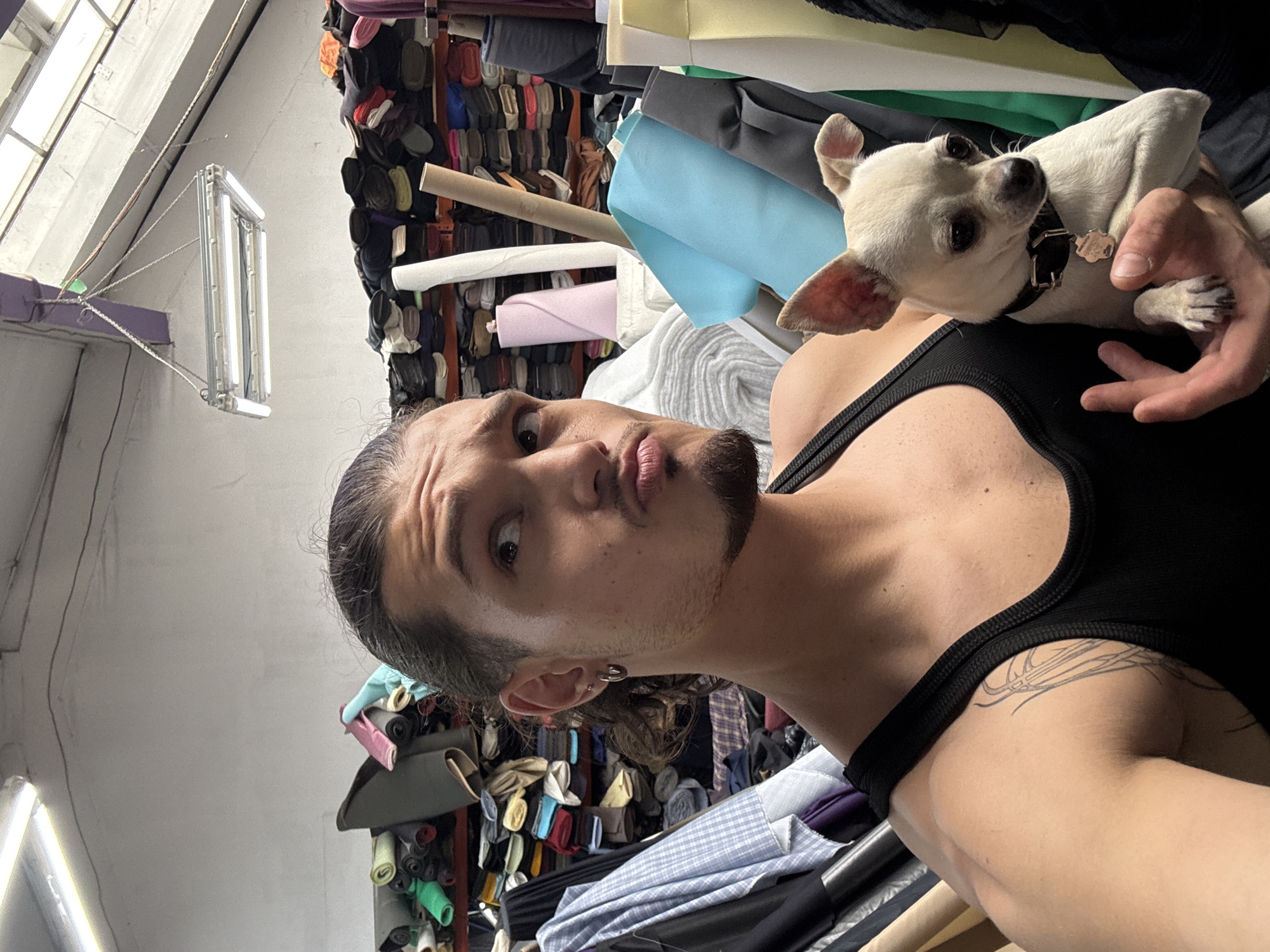 The image features a person holding a small, white dog. The person is looking towards the camera with an open mouth and has dark hair pulled back. They are wearing a black tank top with a tattoo visible on their shoulder. The dog, a chihuahua mix, is held close to the person's chest and is looking away from the camera. They are in a room filled with rolls of fabric and material, suggesting a textile shop or workshop. The background shows shelves stacked high with various colors and textures of fabric, some neatly rolled, others in larger bolts. A fluorescent light fixture is visible hanging from the ceiling. The scene appears to be indoors, with natural light filtering in from a window on the left. The overall mood is informal and personal, like a snapshot taken by the person themselves. The location context indicates this is in Sofia, Bulgaria. No visible text is present in the image.