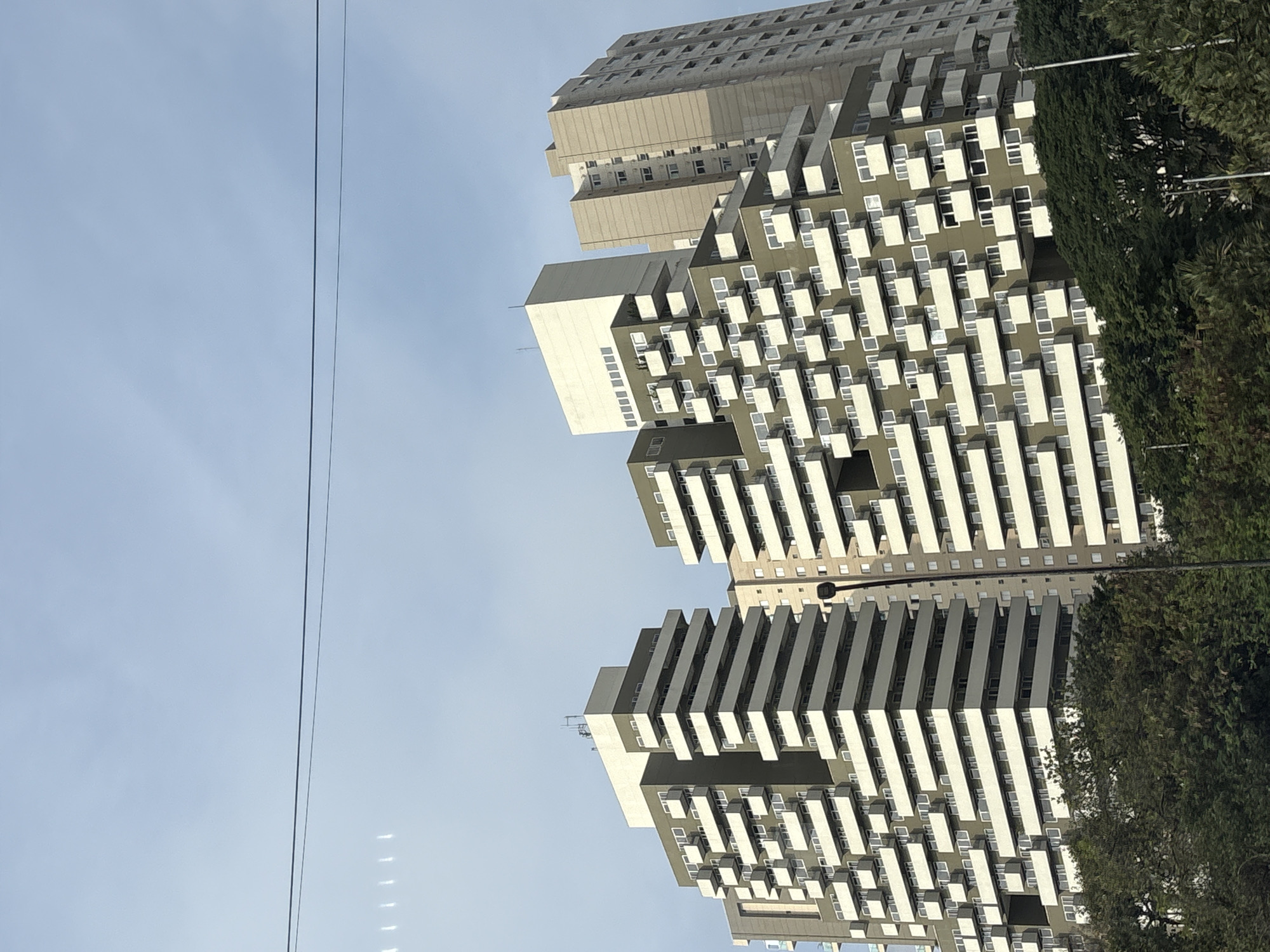 The image shows a cluster of modern high-rise apartment buildings against a pale blue sky with thin, wispy clouds. The buildings feature a stepped, terraced design with numerous balconies. The main building in the foreground is olive green with white balconies and numerous small white-framed windows. To its right, a taller, lighter-colored building is visible, also with many windows. A third, even taller building, light beige in color, is situated to the far right, showcasing a more traditional rectangular facade with repeating rows of windows. Lush green trees and foliage form the base of the scene, obscuring the lower portions of the buildings. Power lines are visible stretching across the upper portion of the sky. The architecture suggests an urban setting, likely a residential area within the city. Given the architectural style and the specific stepped design of the prominent olive green building, this scene is likely located in the city of São Paulo, Brazil, possibly in a district known for its contemporary residential developments. There are no people or active interactions depicted in the image.