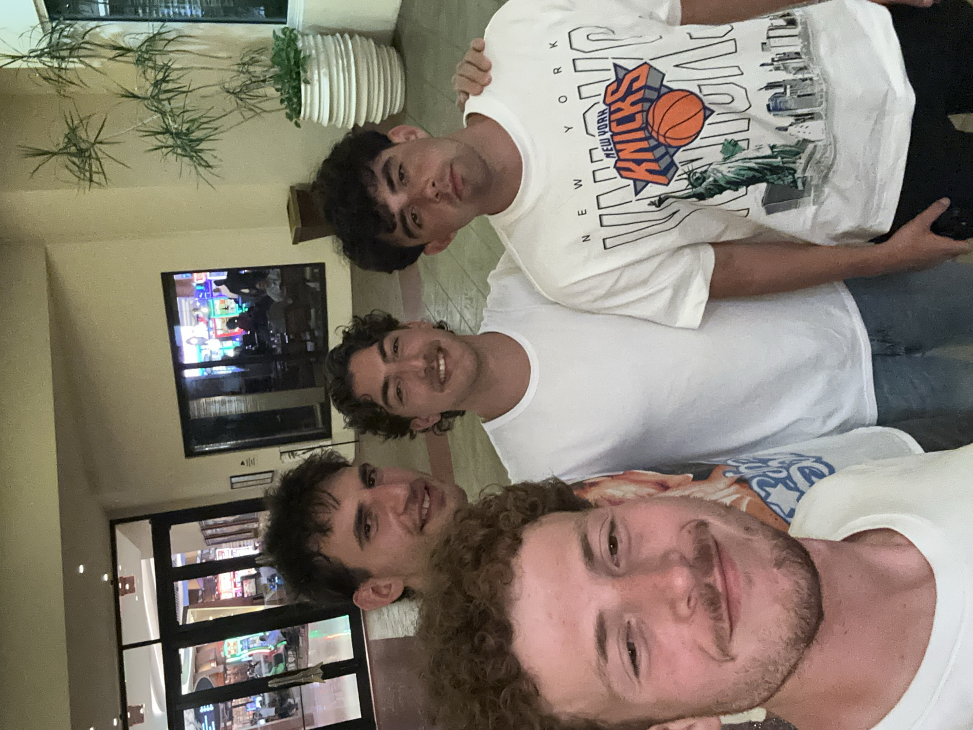 The image shows a group of four young men taking a selfie in what appears to be the lobby of a hotel or resort. The man closest to the camera has curly hair and a light beard. Above him is a man with dark hair and a white t-shirt. Above him is another man with dark curly hair and a white t-shirt. The man at the top is wearing a white New York Knicks T-shirt. The location is likely the Seminole Hard Rock Hotel & Casino Hollywood in Hollywood Seminole Reservation, United States of America, as suggested by the architecture and interior design visible in the background through the glass windows and doorway. A potted plant and a television screen are also visible in the background. They seem to be posing for a casual photo.