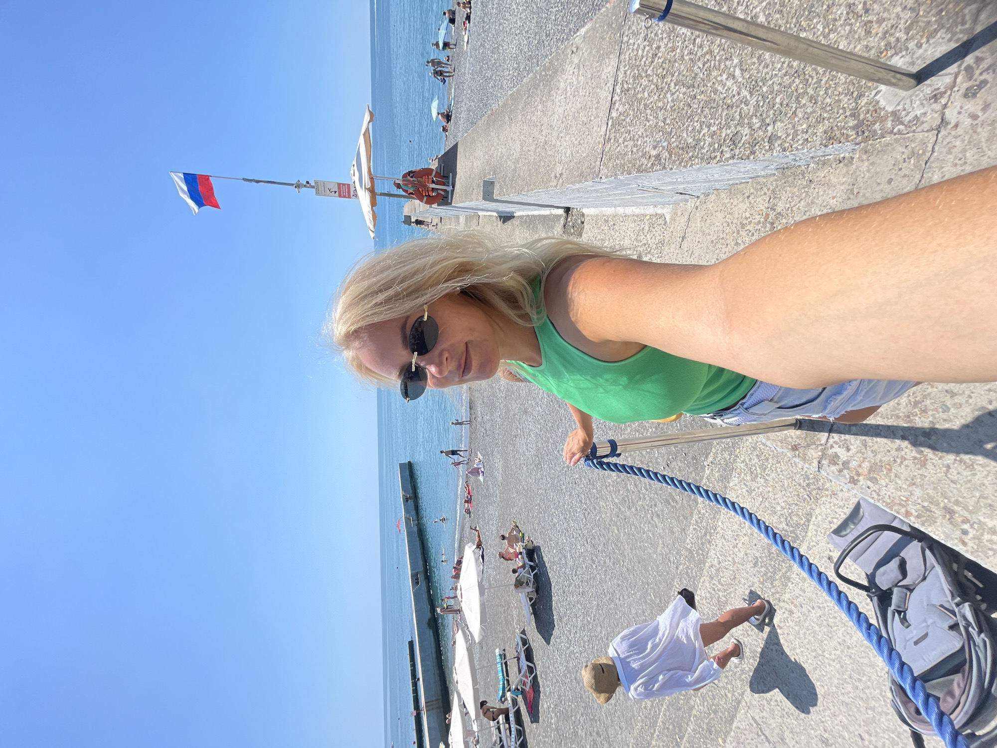 This media file depicts a sunny day at a beach in Sochi, Russia. The scene is set on a pebbly beach with the blue sea stretching out in the background. A woman with blonde hair and sunglasses is taking a selfie, wearing a green tank top and denim shorts. She appears to be enjoying the warm weather.
Several people are visible on the beach, sunbathing on lounge chairs under umbrellas, swimming in the sea, and strolling along the shore. A Russian flag flutters on a pole near a lifeguard station. A stroller is parked on the concrete promenade, and a child in a white dress is walking away from the camera.
The time of day appears to be midday, indicated by the bright sunlight and clear blue sky, suggesting fair weather. The overall mood is one of relaxation and leisure, typical of a beach vacation. The concrete walkway in the foreground is bordered by a metal railing with a thick blue rope.