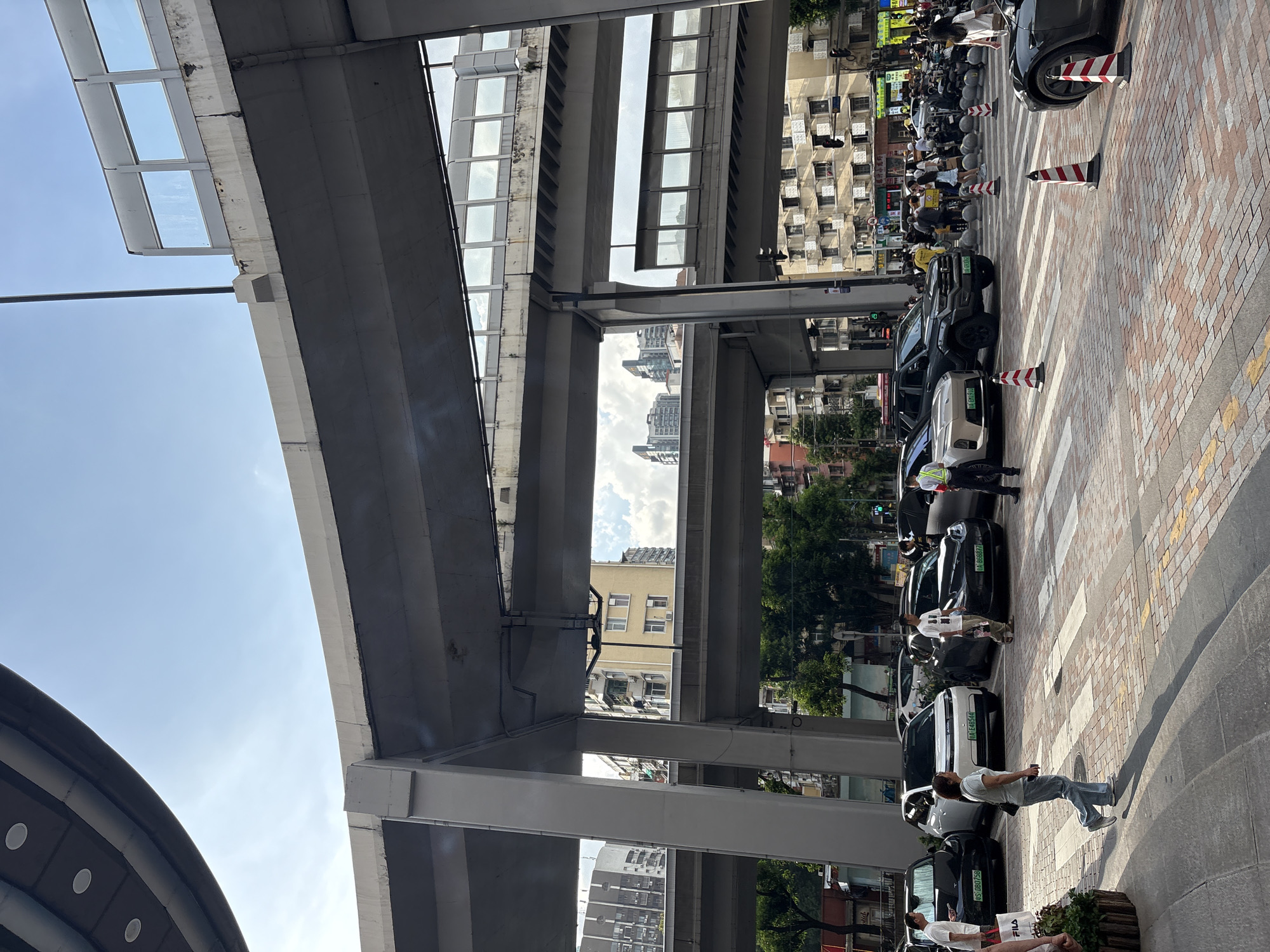 In Haizhu District, China, a pedestrian crossing is marked on a brick-patterned street beneath elevated highways. Several vehicles, including black and white sedans, a dark SUV, and a black sports car, are parked or moving in the vicinity. A group of pedestrians is crossing the street, with some individuals walking and others standing. Traffic cones delineate the pedestrian crossing. In the background, buildings of various heights and architectural styles are visible, with trees and overhead power lines also present. The sky is blue with scattered clouds.