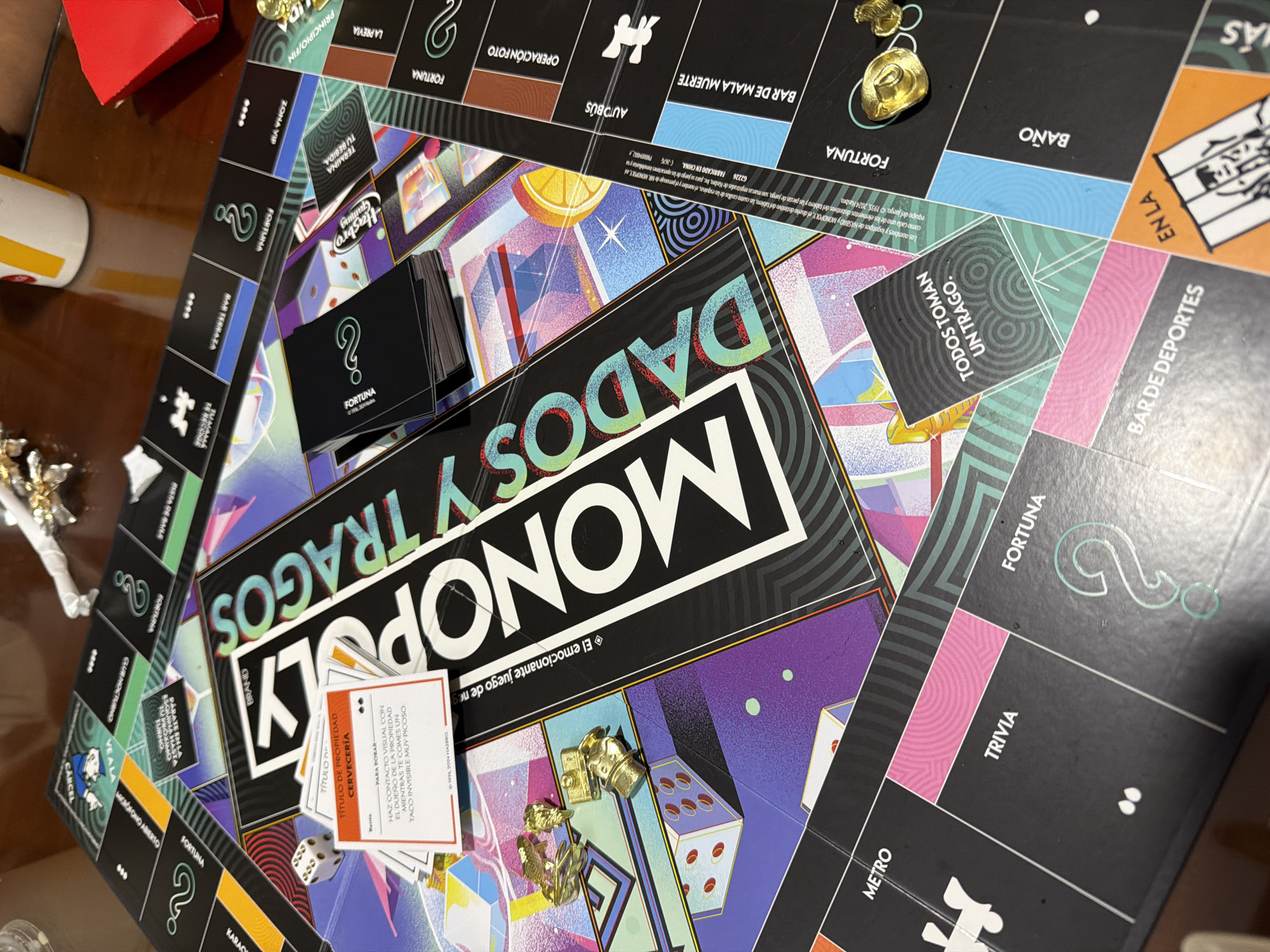 The image shows a close-up of a Monopoly game board, specifically the "Monopoly Dados y Tragos" version. The board is oriented diagonally. The upper left corner contains spaces like "Fortuna," "Zona VIP," and "La Previa." The upper right corner has spaces like "Baño," "En la," and "Bar de Deportes." The lower right corner shows spaces for "Metro" and "Trivia." The lower left corner shows spaces for "Carcel" and "Club Nocturno." The center of the board is largely occupied by the Monopoly game title and logo. Property cards, "Fortuna" cards, game pieces, and dice are scattered across the board.