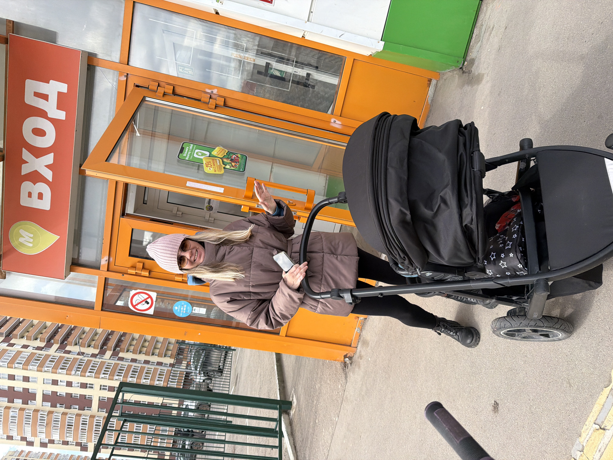 A young woman is captured in a full-length shot, pushing a black baby stroller on a concrete sidewalk in front of a store entrance. She has long blonde hair, is wearing a pink knitted beanie, sunglasses, a brown puffer jacket, black leggings, and black boots. She holds a black smartphone in her right hand and raises her left hand, possibly waving or making a gesture. A cheerful smile is visible on her face.
The storefront features an orange frame with large glass panels and doors. To the left of the entrance, a large red sign with a white border reads "ВХОД" (VKHOD), which means "ENTRANCE" in Russian. Below this text is a green teardrop-shaped logo with a white "M," possibly indicating a supermarket like Magnit. On one of the glass doors, a red circular "No Smoking" sign is visible, along with another green sticker partially advertising "-15%" and text like "Выбирайте" (Choose) and "с собой" (takeaway).
In the background, a large, multi-story residential building with numerous windows and balconies is visible. A green metal railing runs along the foreground, separating the sidewalk from what appears to be a parking or road area. The sky is clear, suggesting a bright daytime setting. The scene evokes a casual outing in an urban environment, likely Perm, Russia, given the provided context.