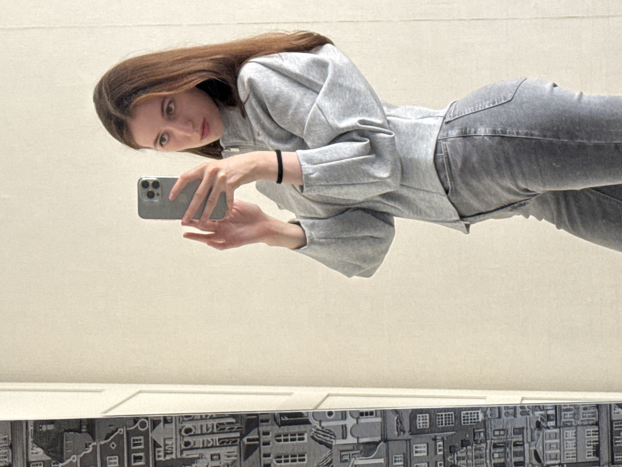 A young woman with long, brown hair takes a mirror selfie. She is wearing a light grey hooded sweatshirt with voluminous sleeves and grey jeans. Her left hand holds a silver smartphone, angled to capture her reflection. A black elastic band is visible on her left wrist. The background is a plain, light-colored wall, with a decorative mural or detailed picture frame visible at the bottom of the image, depicting a cityscape with many buildings and windows. The lighting suggests an indoor setting. The woman's expression is curious and direct, as she looks at the camera.