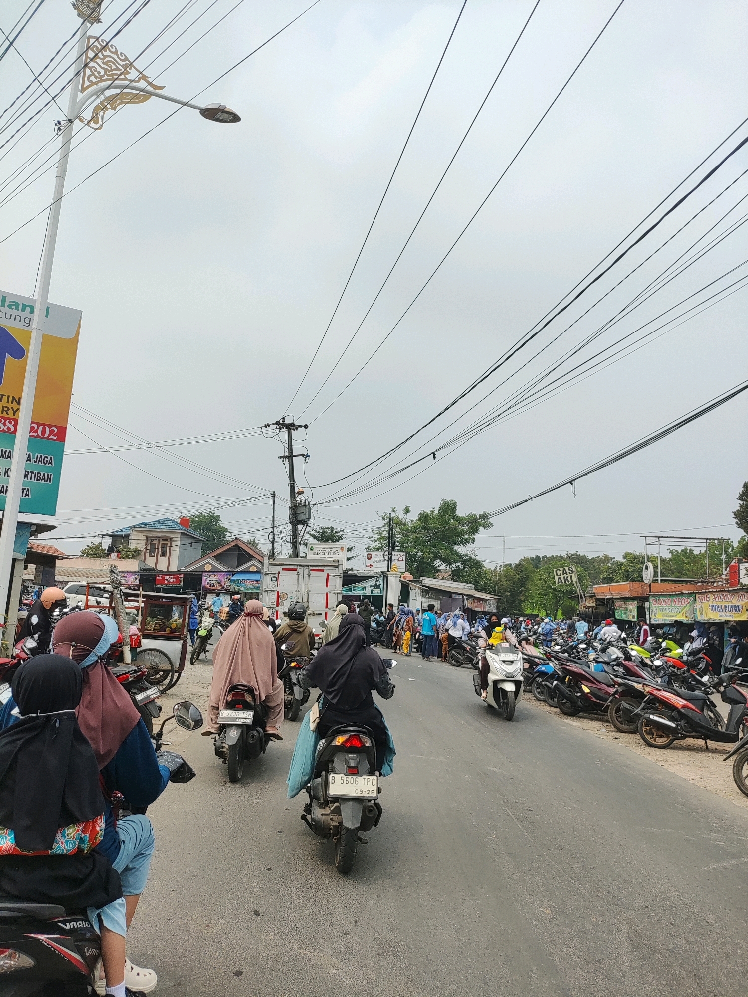 The image depicts a busy street scene in Wanajaya, Indonesia, on an overcast day. The road is filled with numerous motorcycles and scooters, with many people riding them, some wearing helmets and others with headscarves. The traffic appears to be moving slowly, suggesting congestion. On the sides of the road, there are various businesses and stalls, including what looks like a food vendor with a cart, and several shops with signage in Indonesian. The background shows a mix of buildings, trees, and utility poles with a dense network of electrical wires crisscrossing the sky. The overall atmosphere suggests a typical urban environment with a high volume of motorcycle transportation.
