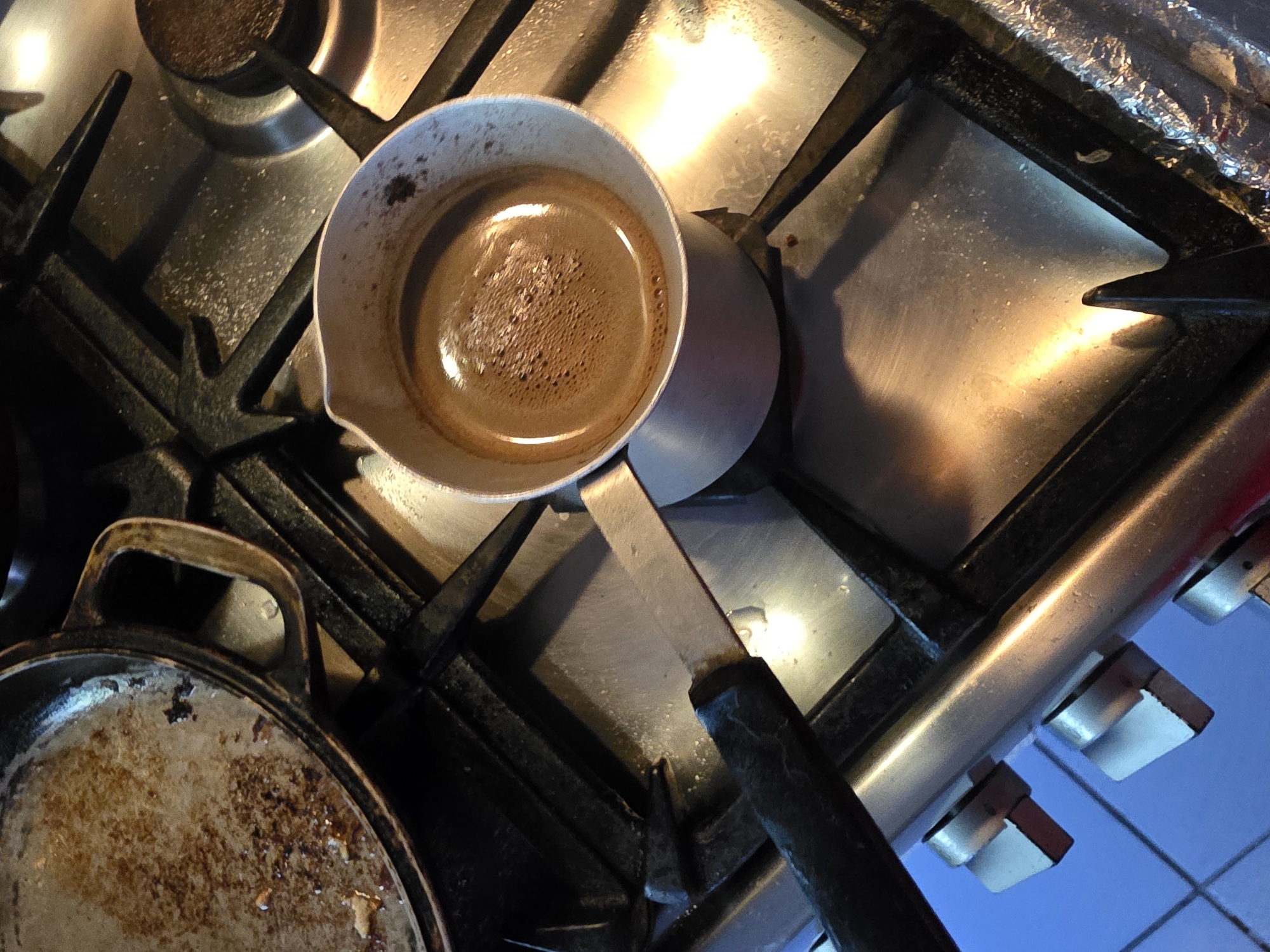 A stovetop in an indoor kitchen in Tuapse, Russia, shows a partially visible old cast iron pan with remnants of food in it, and a small metal pot filled with dark liquid, likely coffee. The pot has a black handle and is placed on the gas burner. The stovetop appears to be made of stainless steel and is showing signs of use and some light residue. The lighting suggests it might be daytime, with artificial light also contributing to the reflections on the metal surfaces. The overall impression is a utilitarian kitchen environment.