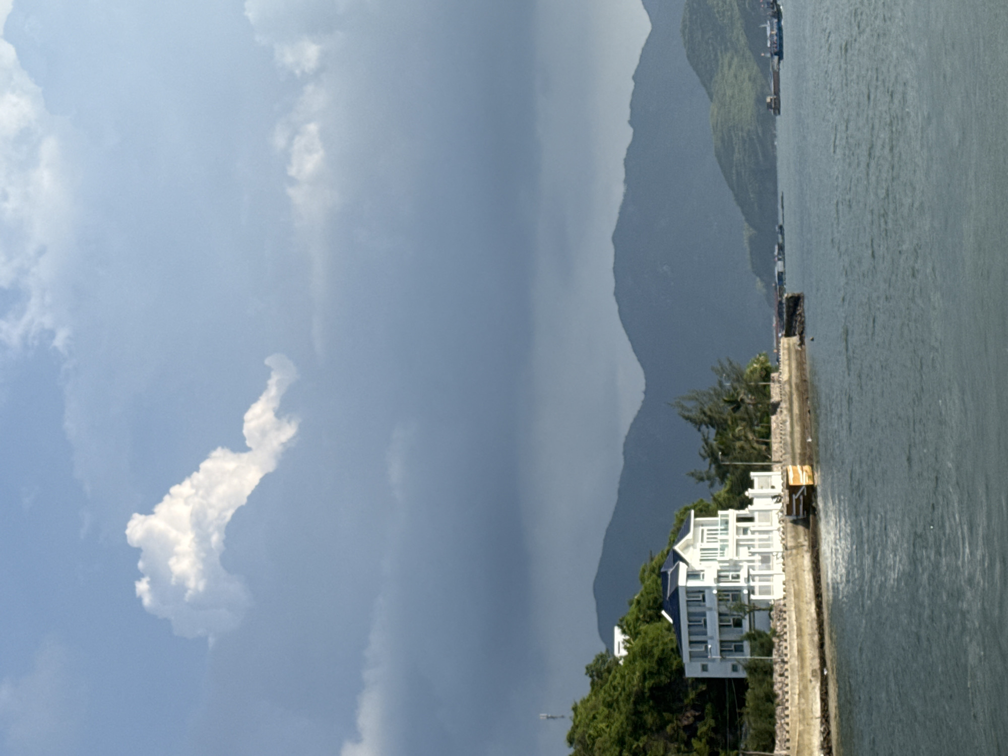The image depicts a coastal scene in Nha Trang Ward, Vietnam. In the foreground, the turquoise waters of the ocean stretch towards the horizon. A multi-story white building with a blue-tiled roof sits prominently on the shore, surrounded by lush green vegetation and a stone wall. The building appears to be a residence or a small hotel, with balconies and large windows. Beyond the building, a series of rolling green hills form the backdrop, their peaks fading into the misty sky. The sky itself is a mix of bright blue and dark, dramatic clouds, suggesting a dynamic weather system. In the distance, several boats are visible on the water, hinting at maritime activity in the area. The overall atmosphere is serene and picturesque, characteristic of a tropical coastal landscape.