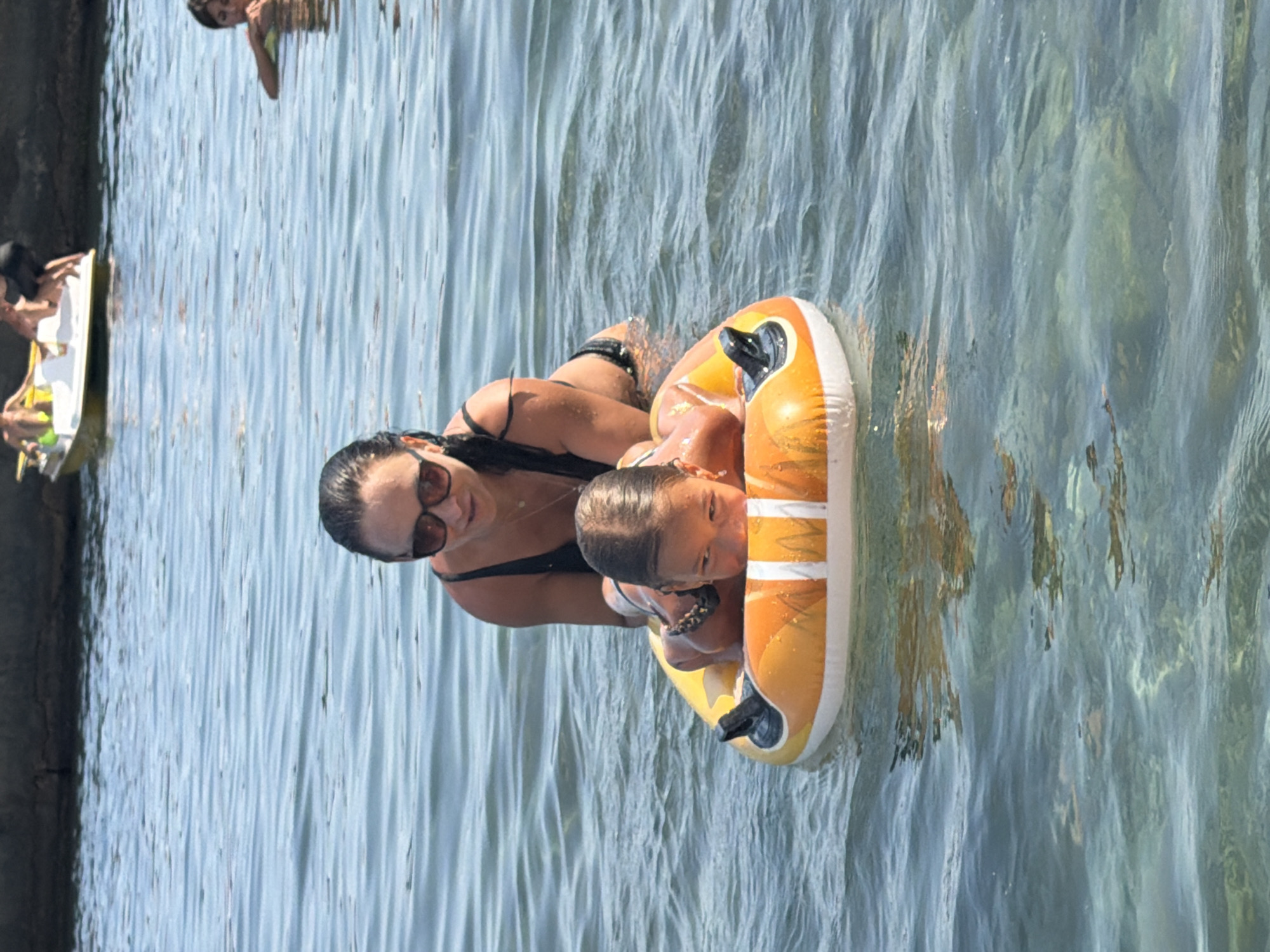 In a bright, sunlit scene, two individuals are enjoying a day in the water. A woman, wearing a black bikini and sunglasses, is holding onto an inflatable ring. A young child, likely a toddler, is nestled within the ring, looking towards the camera with a bright expression. The water is clear and shallow, revealing the sandy bottom and scattered rocks. The reflection of sunlight creates a shimmering effect on the water's surface.
In the background, another person can be seen on a yellow and white board, possibly a paddleboard or a small raft. The setting appears to be a beach or a shallow bay, with a dark, textured wall on the left side of the frame, suggesting a man-made structure or a rocky embankment. The overall mood is one of leisure and fun, with the clear water and bright sunlight indicating a pleasant, warm day. There is no visible text in the image.