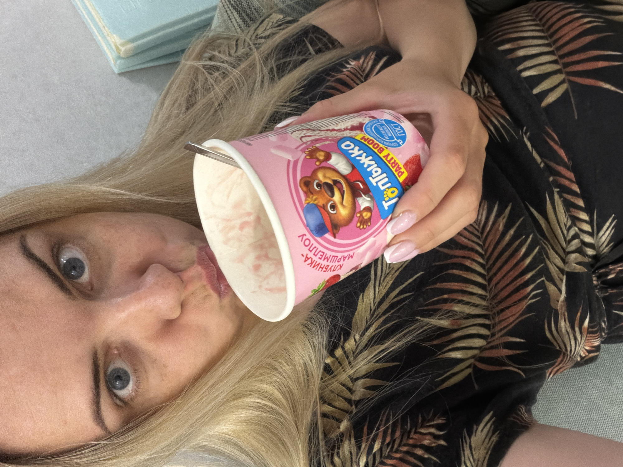A woman is lying down while holding a pink yogurt cup with a cartoon bear on it. She appears to be tasting the yogurt. The background includes part of a couch and a stack of light blue books. The scene is indoors, but there's no specific indication of the city of Votkinsk, Russia.